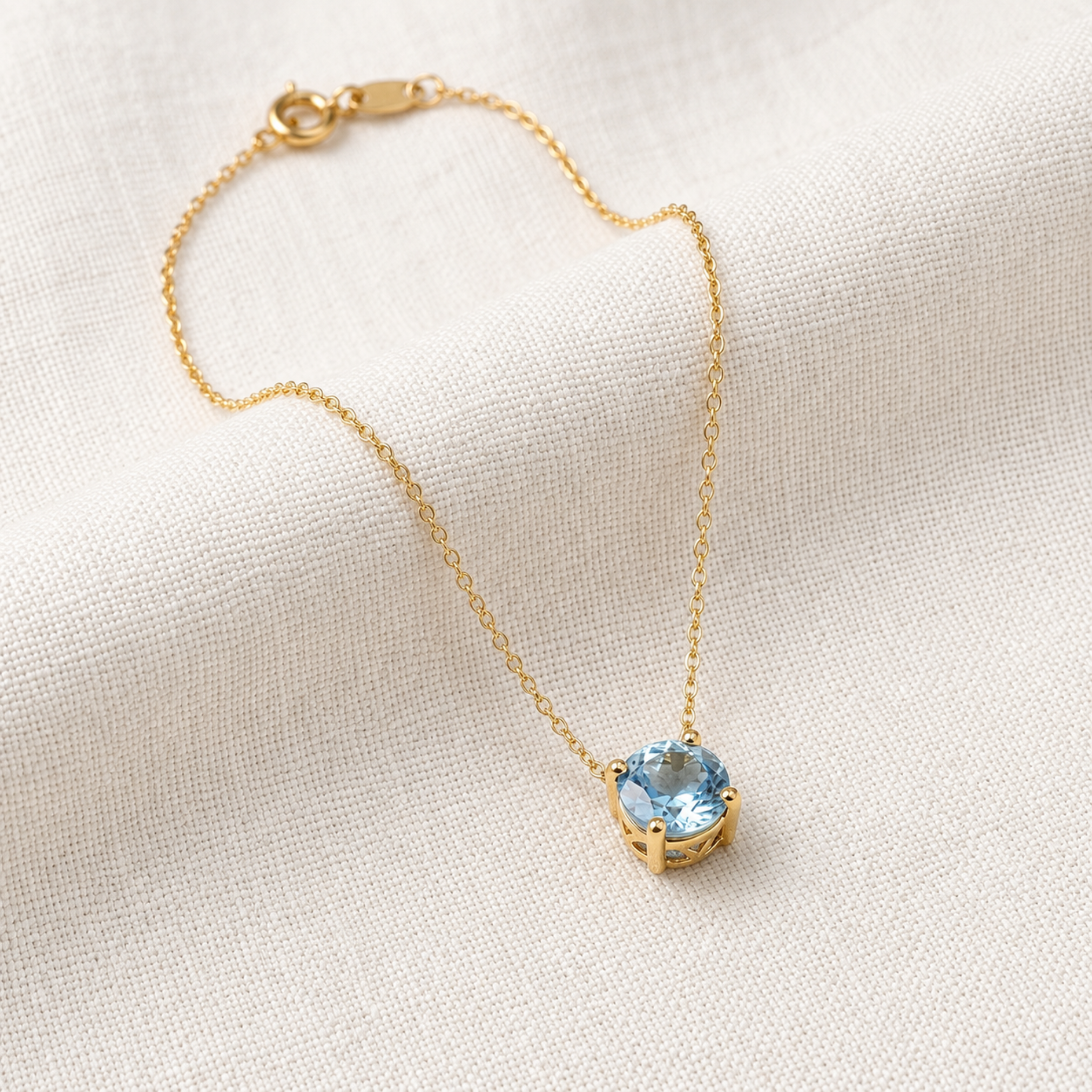 A delicate gold chain necklace with a round bezel-set light blue gemstone pendant, displayed on a textured off-white fabric background.