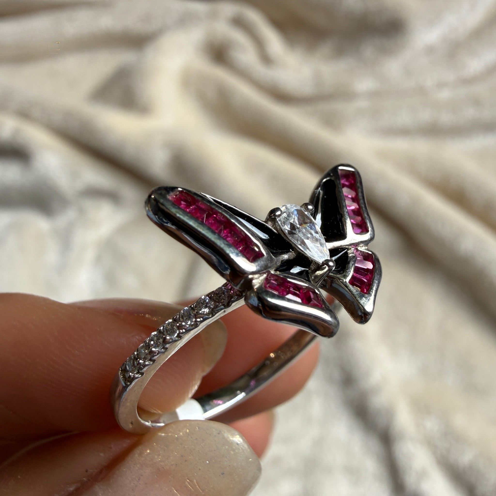 A silver butterfly ring with pink gemstone accents and a clear center stone, held between fingers against a soft beige fabric background.