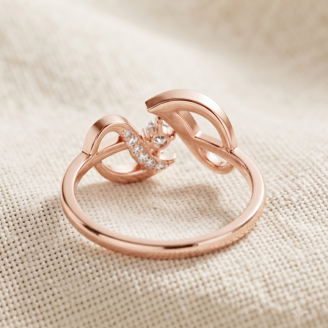A rose gold ring with a stylized infinity symbol design, featuring a small cluster of clear pavé stones on one side, displayed on a textured beige fabric background.