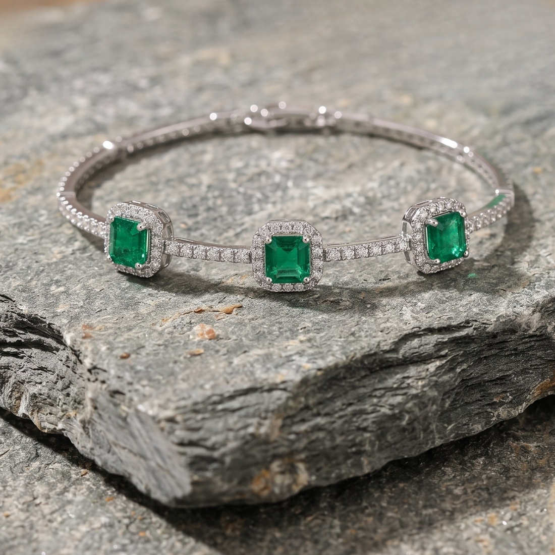 A silver-tone three-stone bangle bracelet featuring emerald-cut green gemstones and pavé-set clear stones, displayed on a textured gray rock.
