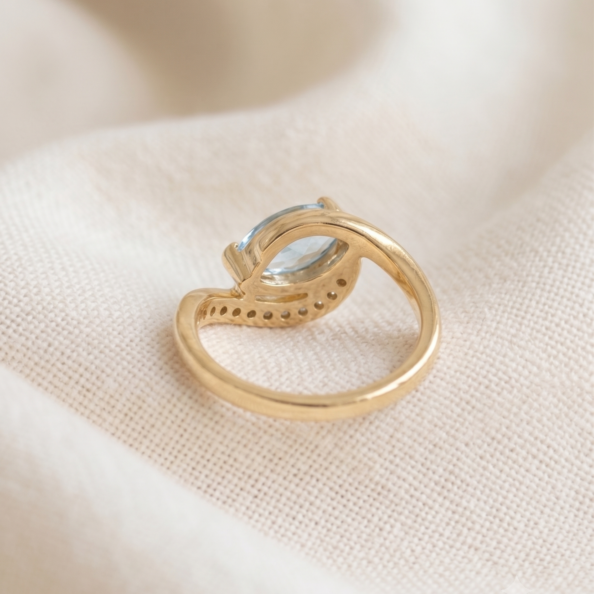 A gold ring with a curved, openwork band and a small oval light blue gemstone, resting on a textured cream fabric background.