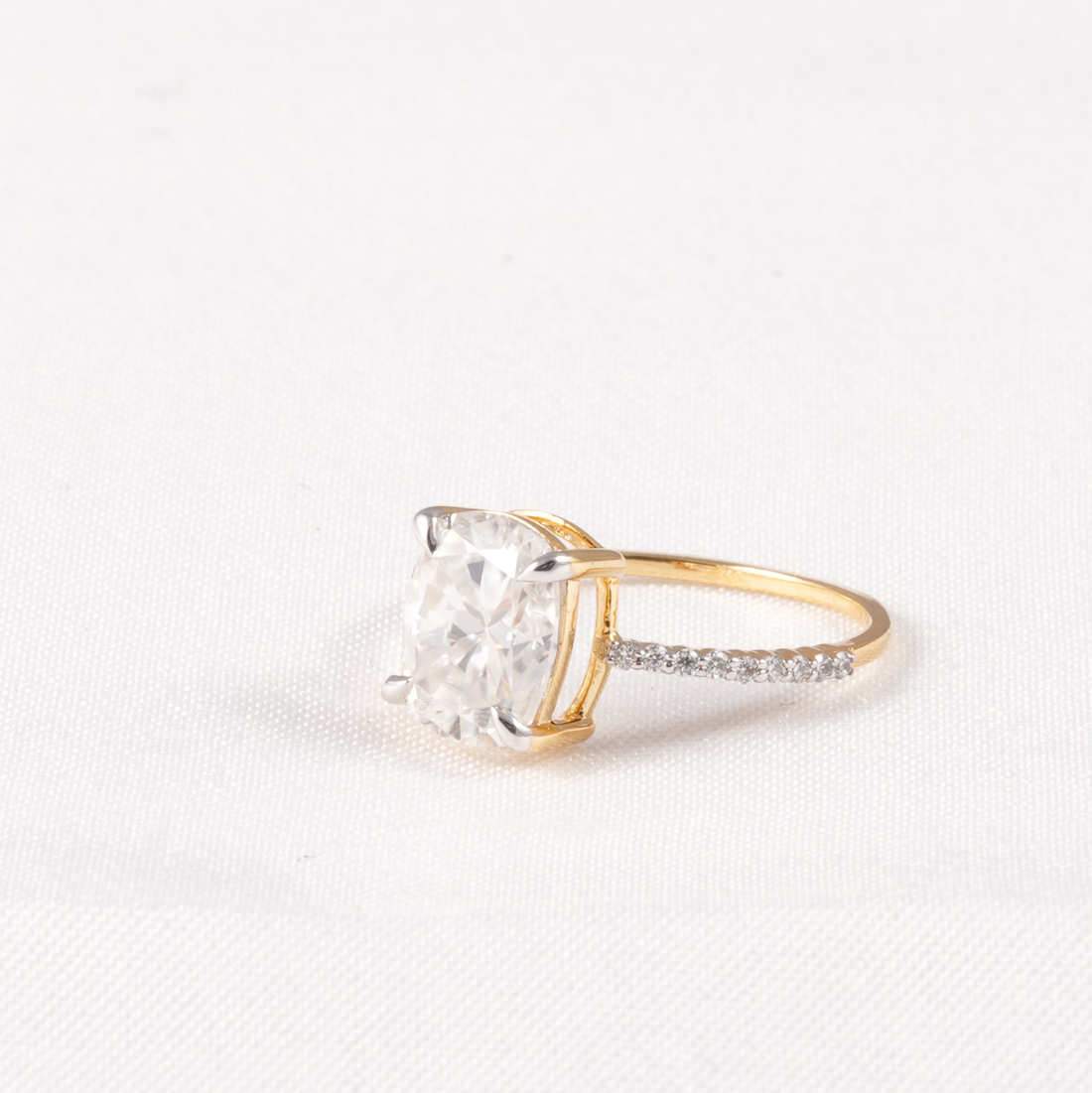 A yellow gold solitaire ring featuring a large cushion-cut clear gemstone in a four-prong setting, with a pavé band accented by small round stones, shown on a textured white background.