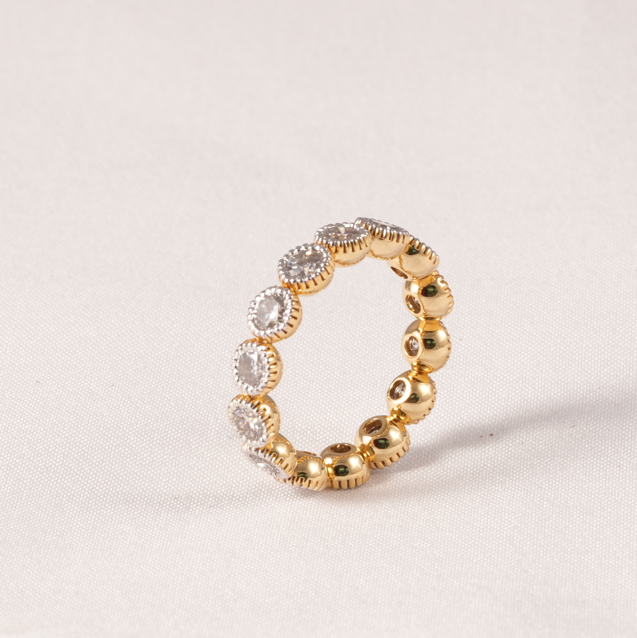 A gold eternity ring featuring a row of bezel-set clear round gemstones, photographed on a neutral textured light beige background.