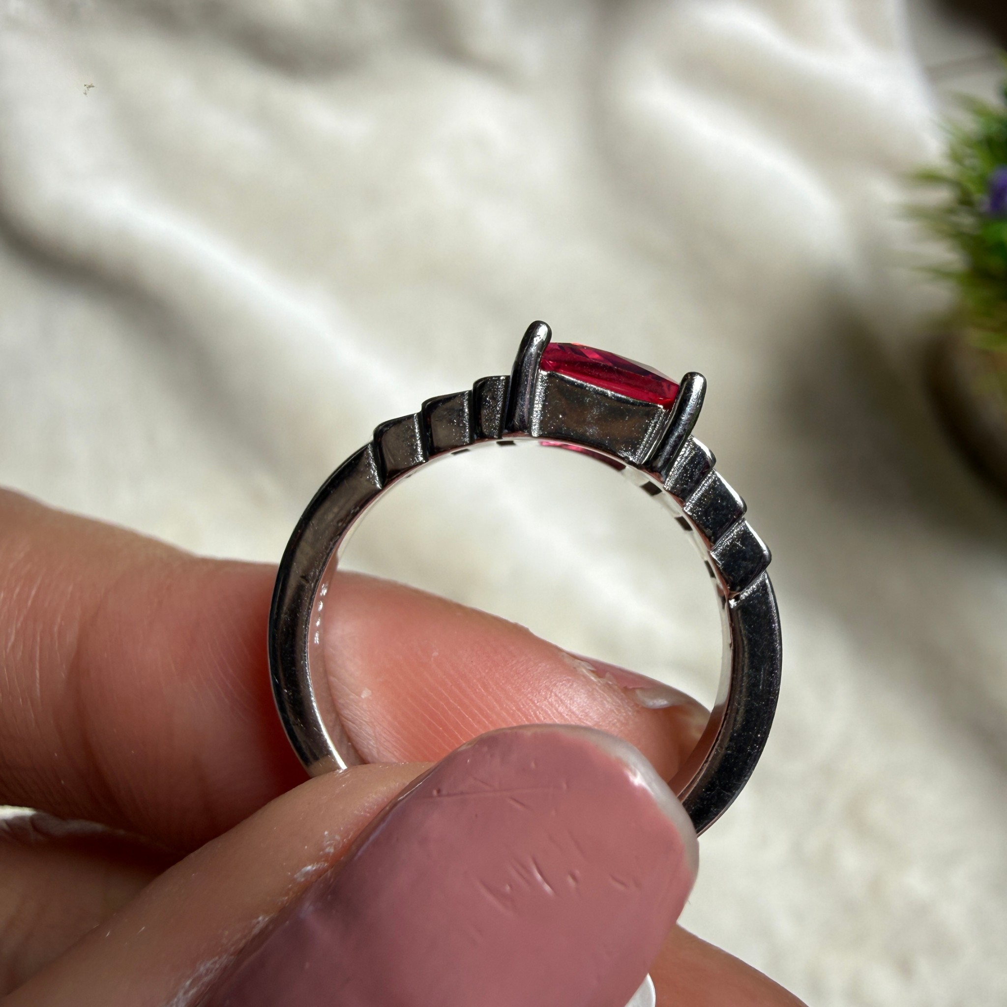 A close-up of a silver ring with a rectangular red gemstone held between fingers against a soft white fabric background.