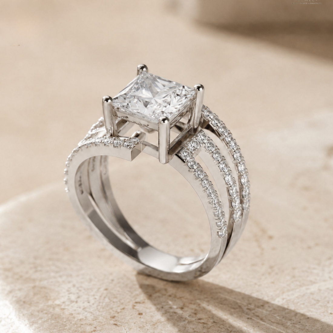 A silver-toned diamond ring featuring a princess-cut clear center stone and pavé-set accent diamonds along curved split shanks, photographed on a warm beige stone surface.