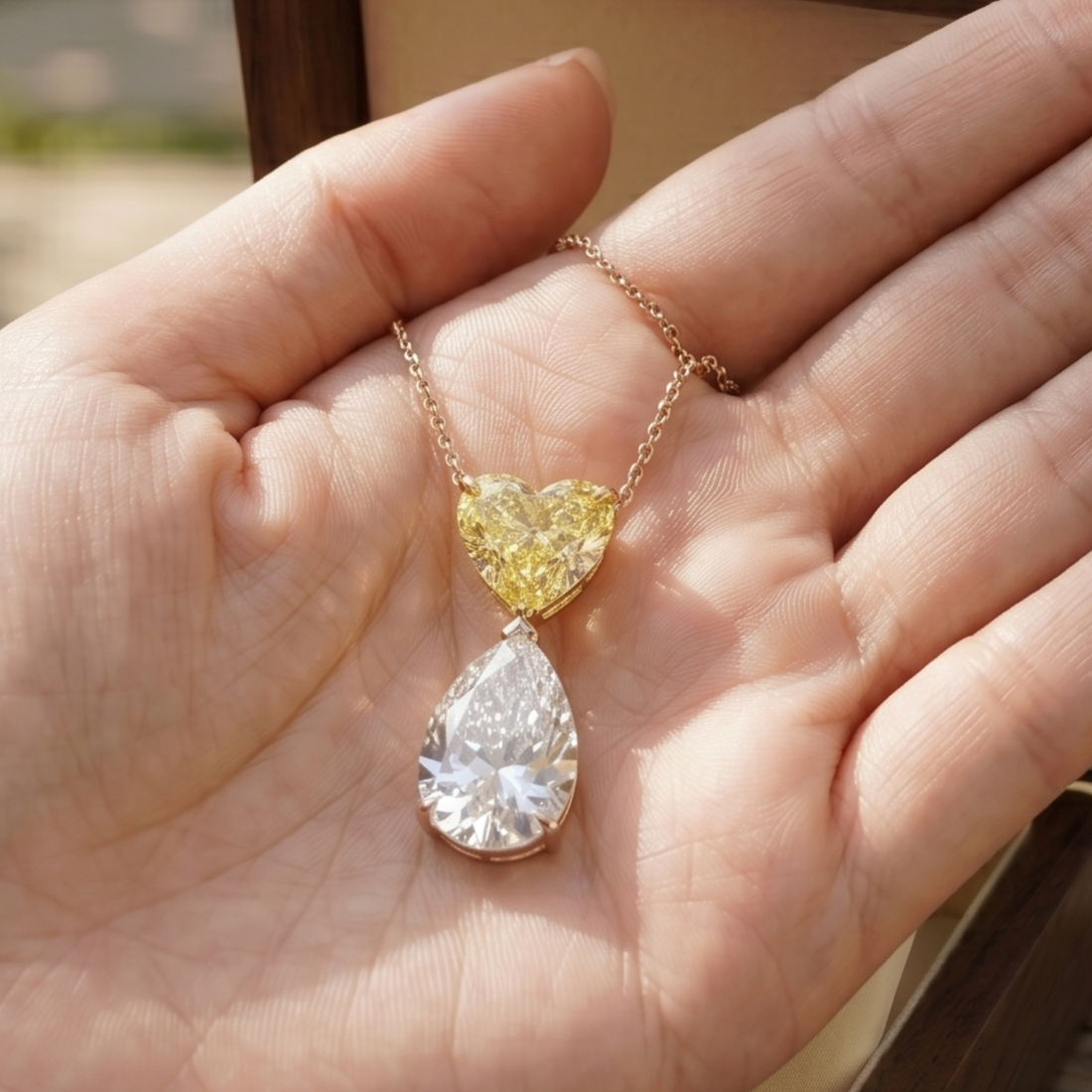 A gold necklace with a heart-shaped yellow gemstone and a teardrop-shaped clear gemstone pendant, held in a person's open palm.