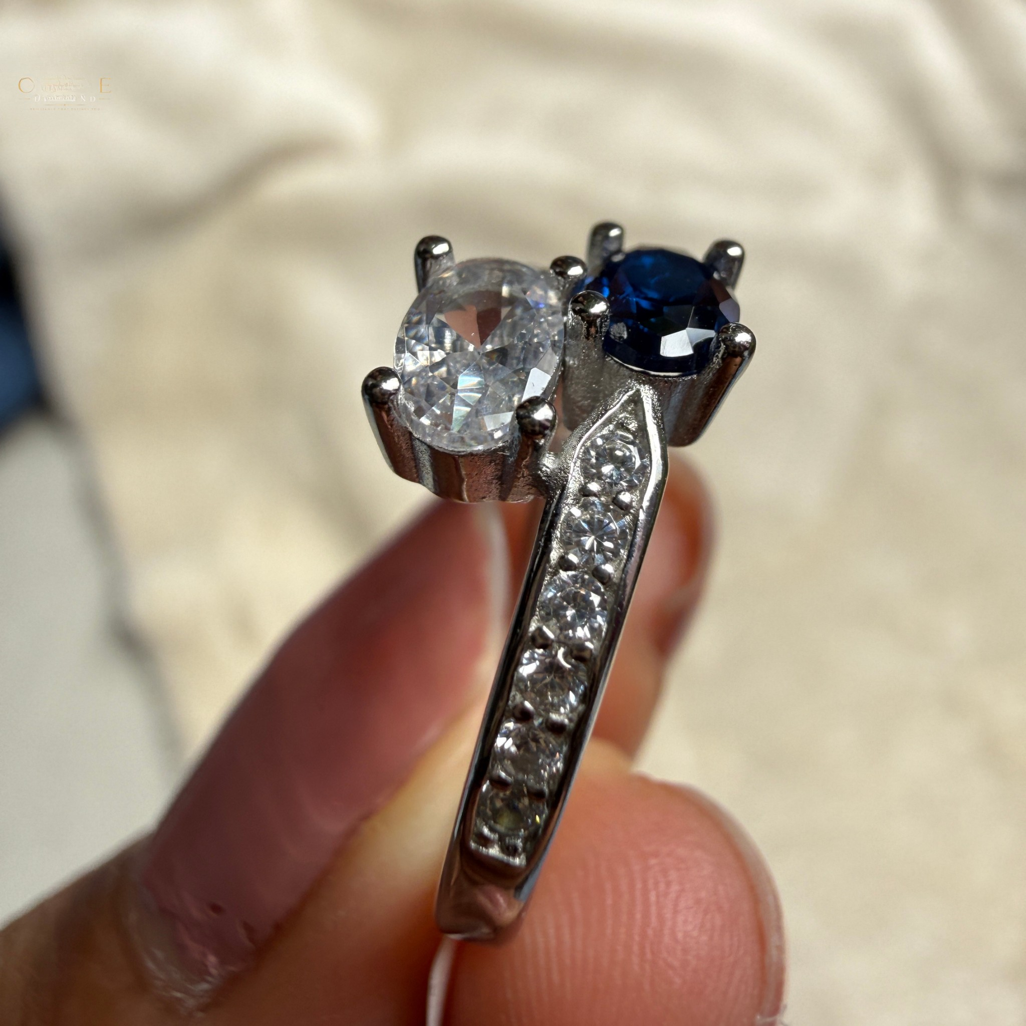 A close-up of a silver-toned ring held between fingers, featuring a large round clear center stone and a smaller round dark blue stone, with a pavé-set row of small clear stones along the band.