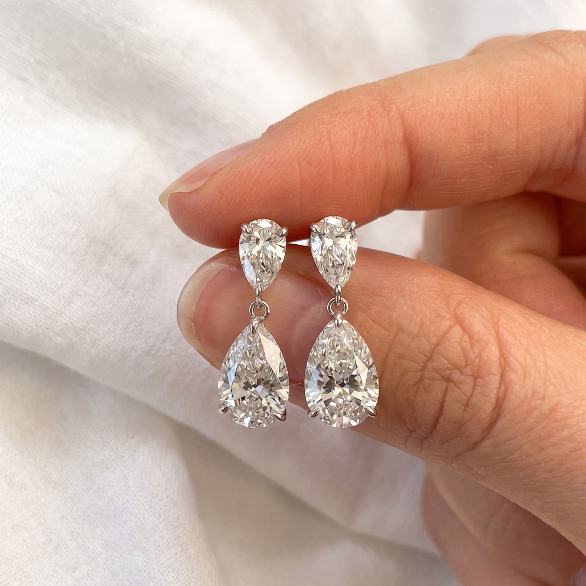 A pair of pear-cut diamond drop earrings in silver-toned settings, held between fingers against a soft white fabric background.
