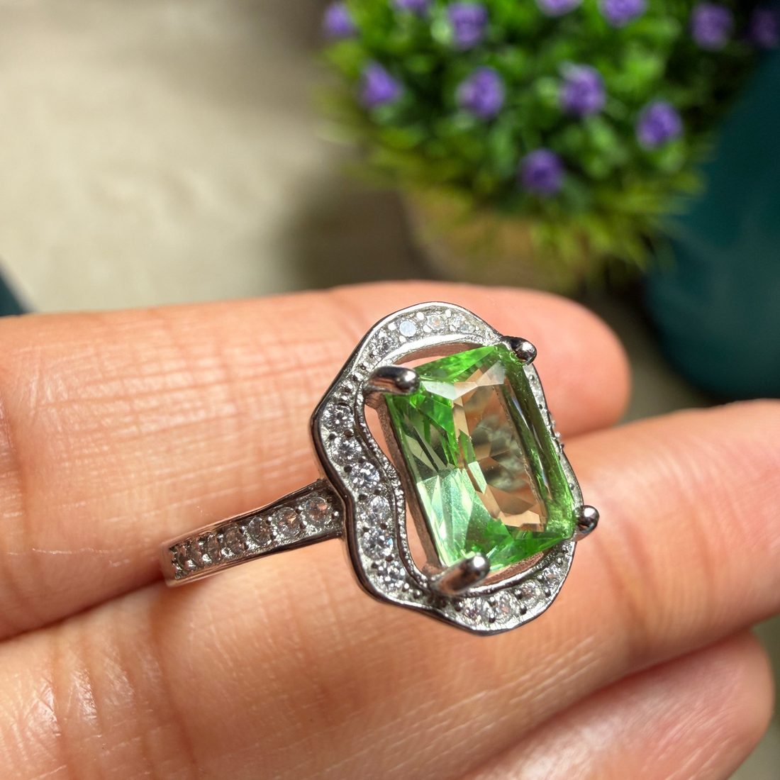 A silver ring with a rectangular green gemstone and small clear accent stones, shown resting on a person's fingers with a blurred background of greenery and purple flowers.