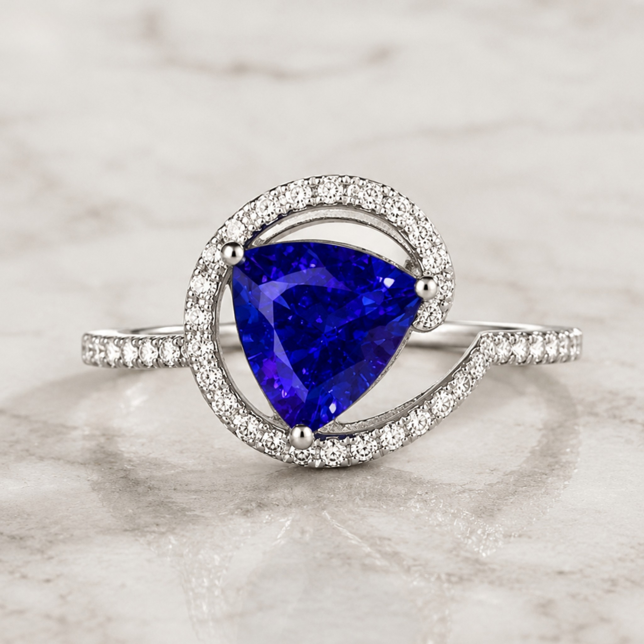 A silver ring featuring a large, brilliant-cut deep blue triangular gemstone, surrounded by a double halo of small clear pavé stones, set on a polished silver band with pavé accents, displayed on a light marble surface.
