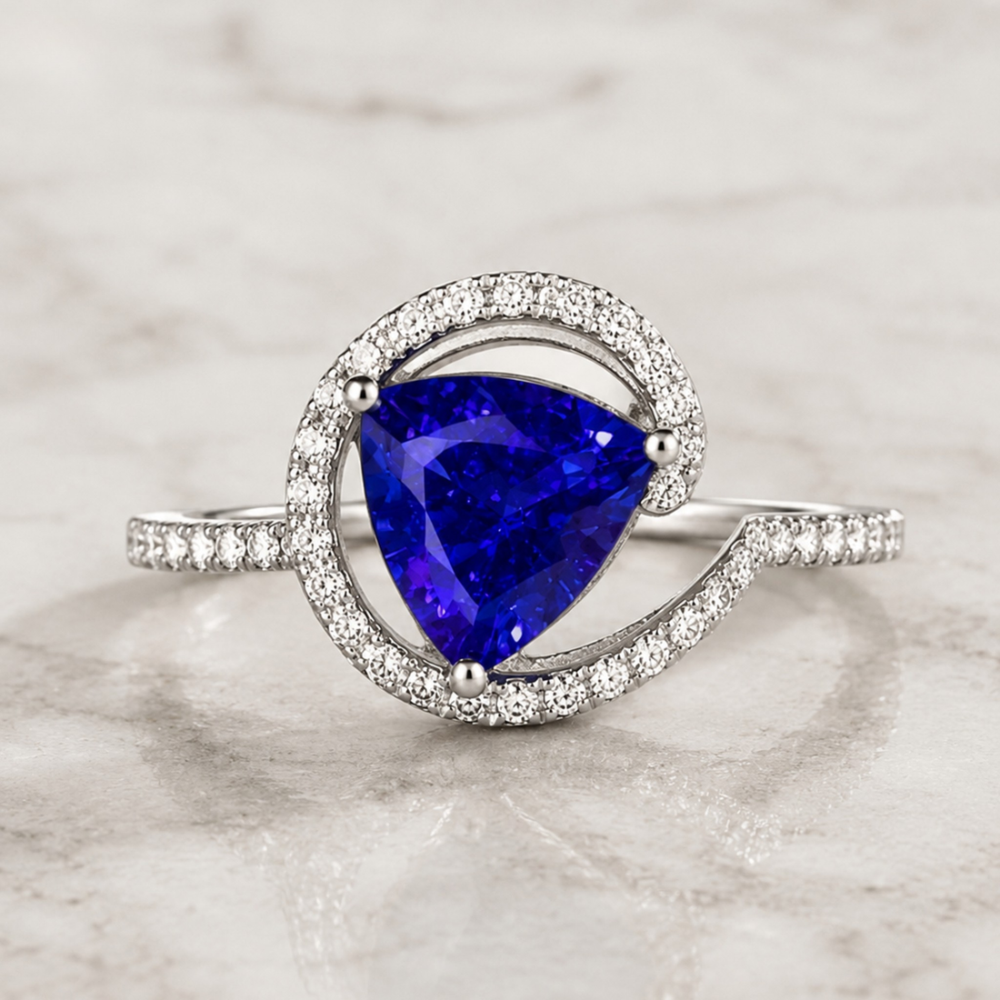 A silver ring featuring a large, brilliant-cut deep blue triangular gemstone, surrounded by a double halo of small clear pavé stones, set on a polished silver band with pavé accents, displayed on a light marble surface.