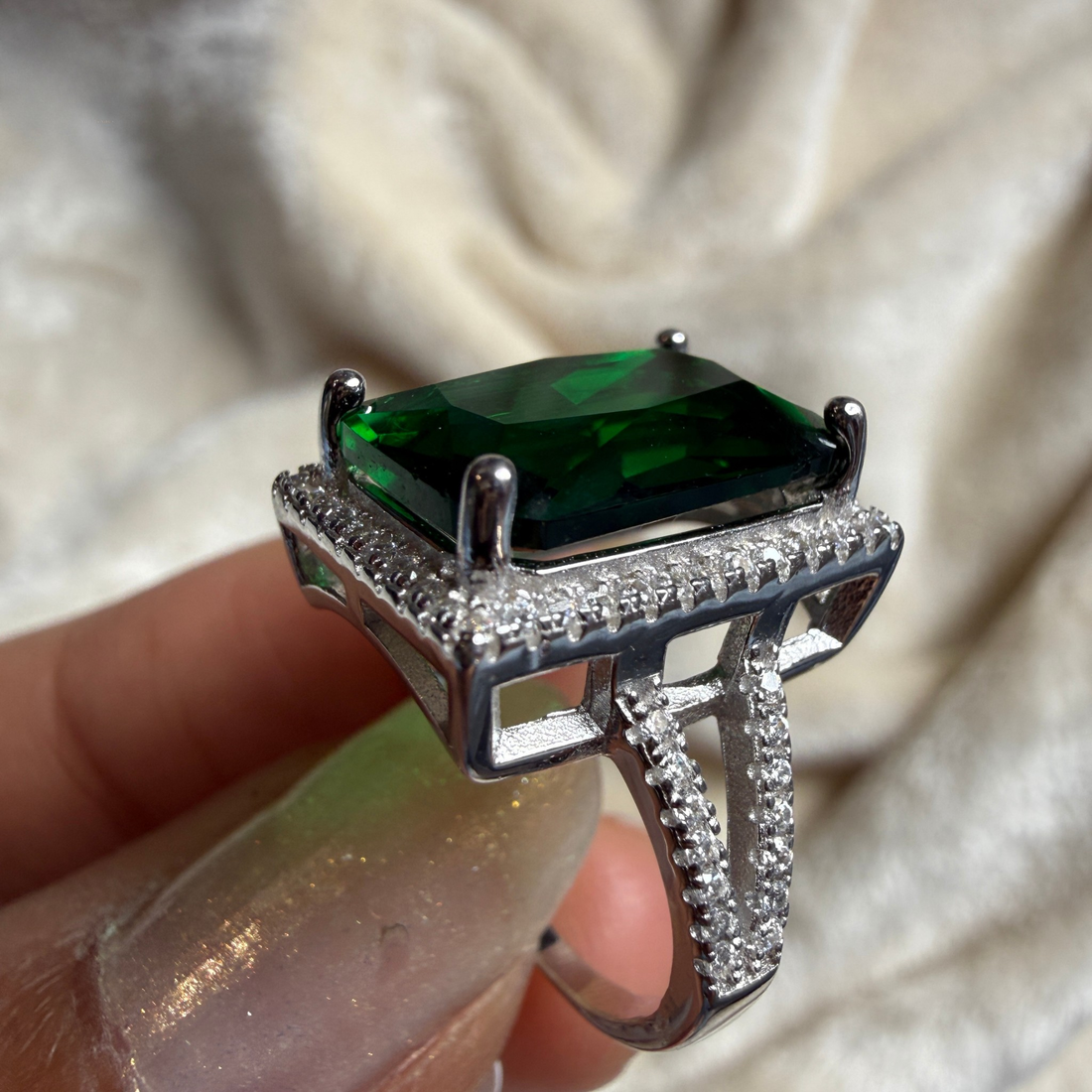 A silver ring with a large rectangular green gemstone and small clear stones, shown being held on a finger against a soft cream fabric background.