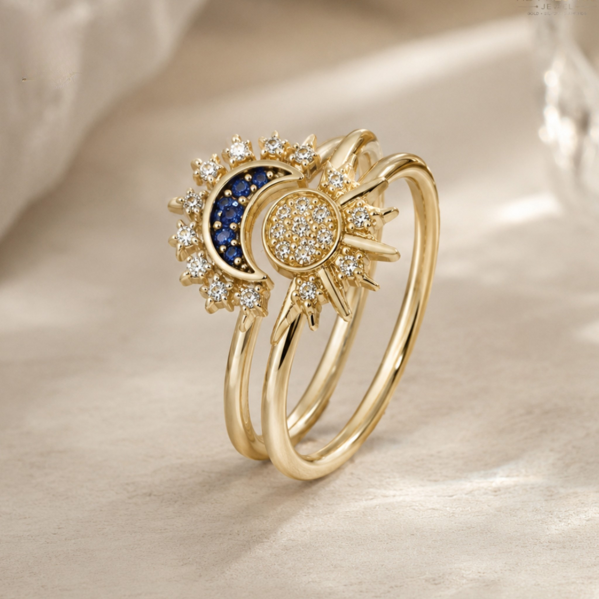 A gold double-band ring featuring a sun and moon design, with a dark blue crescent moon, a gold sun face paved with small clear stones, and surrounding gold sunburst details, set on a soft beige fabric background.