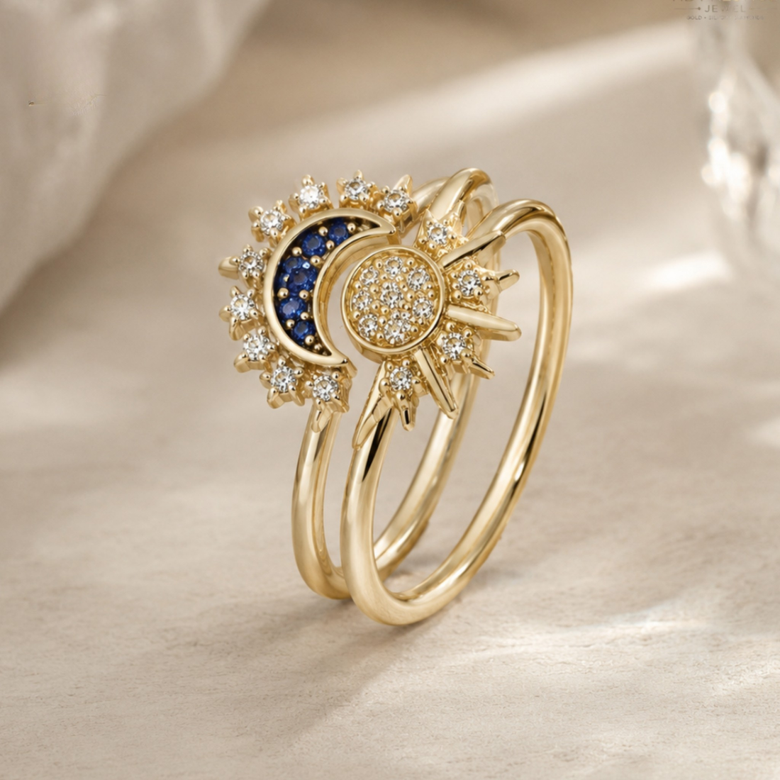 A gold double-band ring featuring a sun and moon design, with a dark blue crescent moon, a gold sun face paved with small clear stones, and surrounding gold sunburst details, set on a soft beige fabric background.