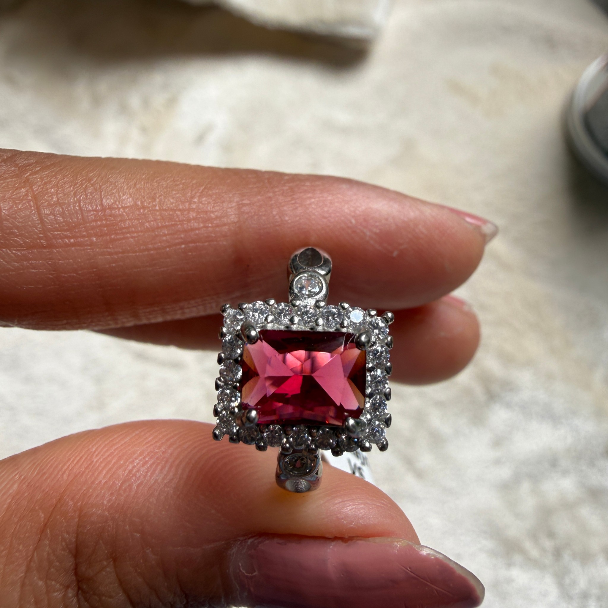 A silver-toned ring with a rectangular red gemstone surrounded by small clear stones, held between fingers against a soft beige background.
