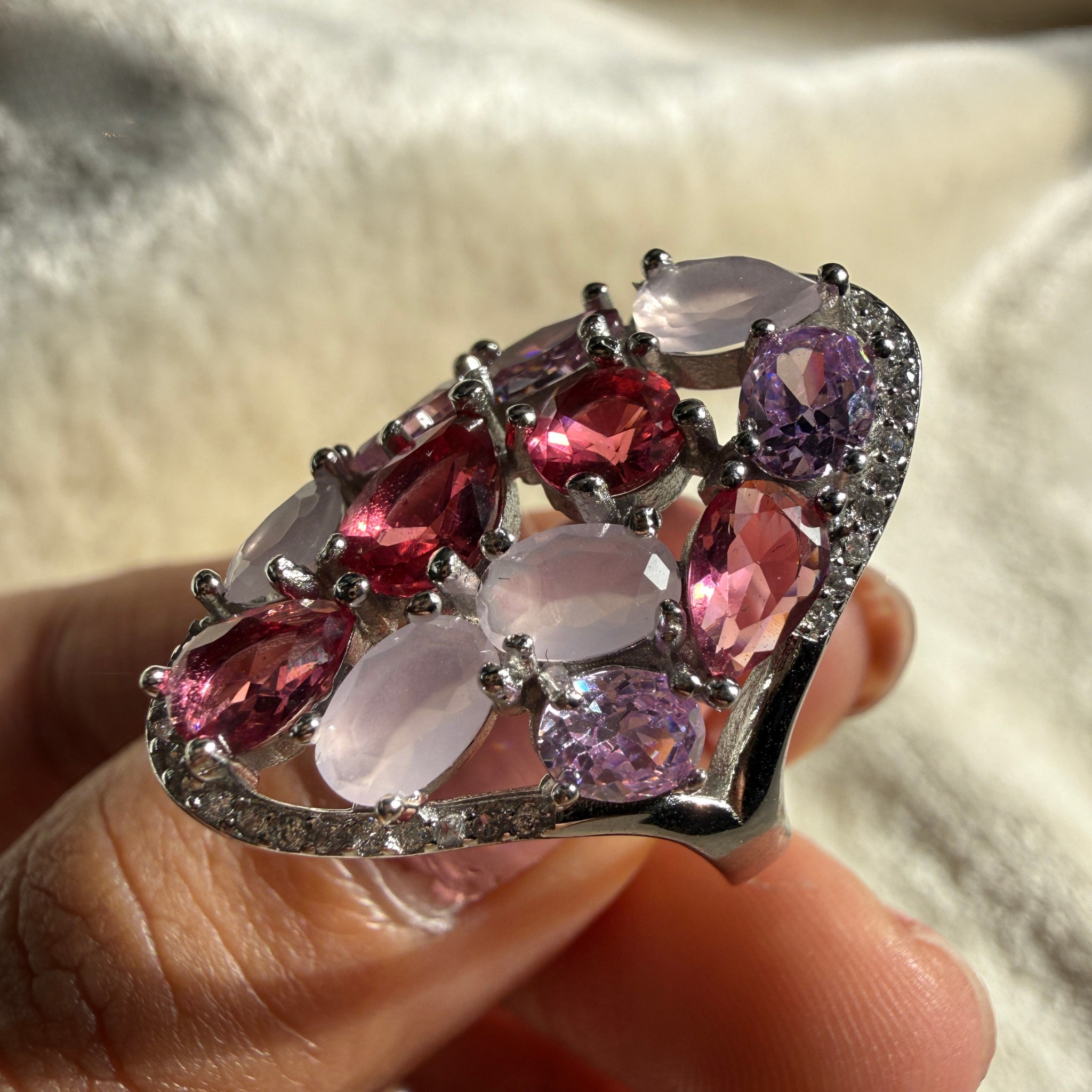 A close-up of a silver-toned ring held on a person's fingers, featuring multiple oval-cut pink and purple gemstones with small clear accent stones along the band, set against a soft cream fabric background.