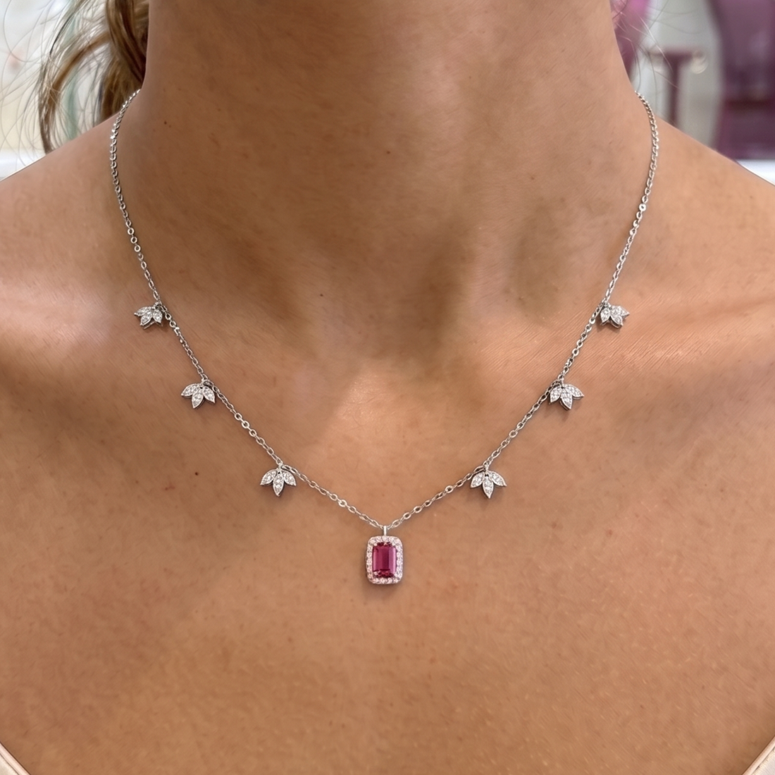 A silver-toned delicate necklace with five small leaf-shaped pavé settings and a rectangular emerald-cut pink gemstone pendant, shown worn on a model's neck.
