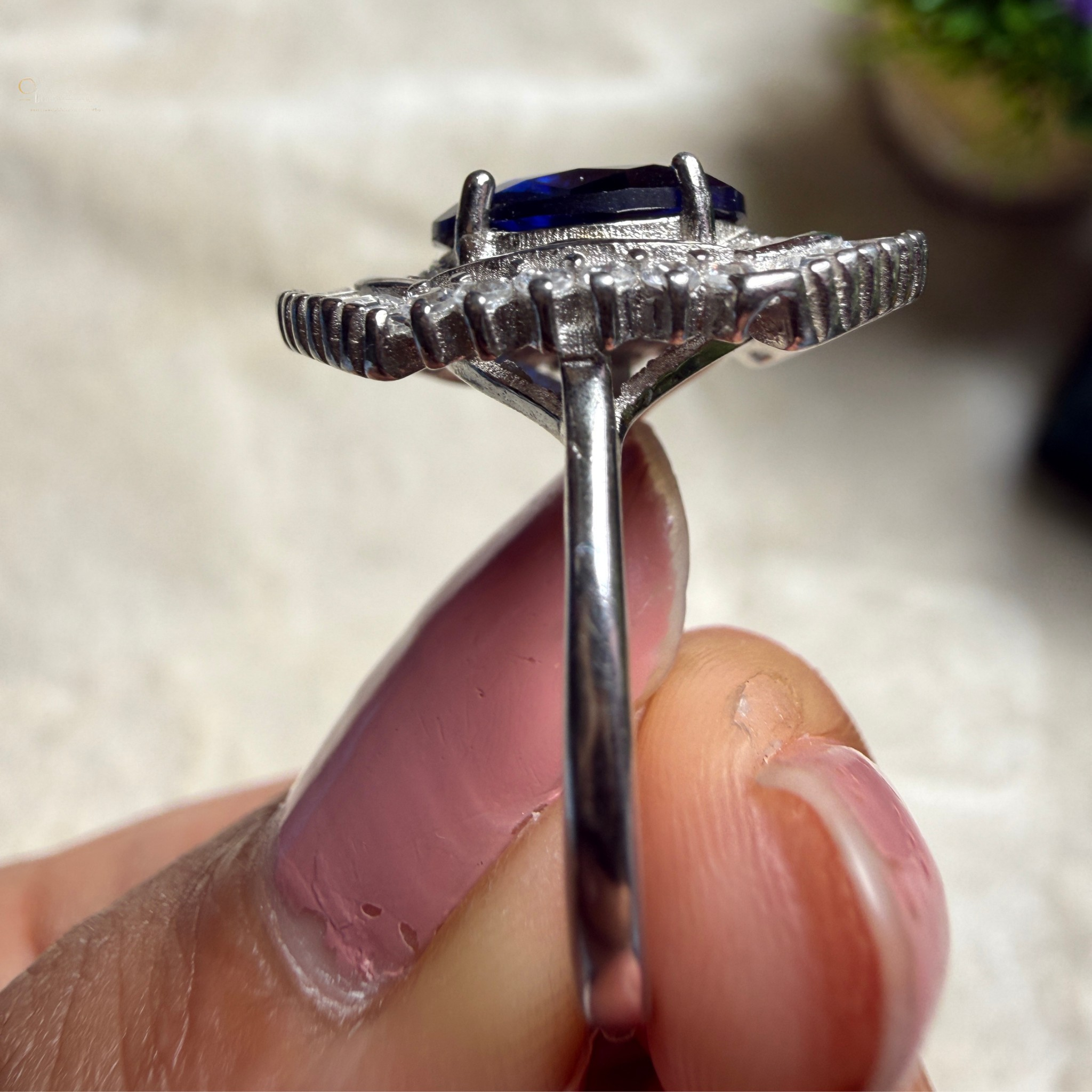 A close-up of a silver ring with a dark blue oval gemstone, held between fingers against a soft beige background.