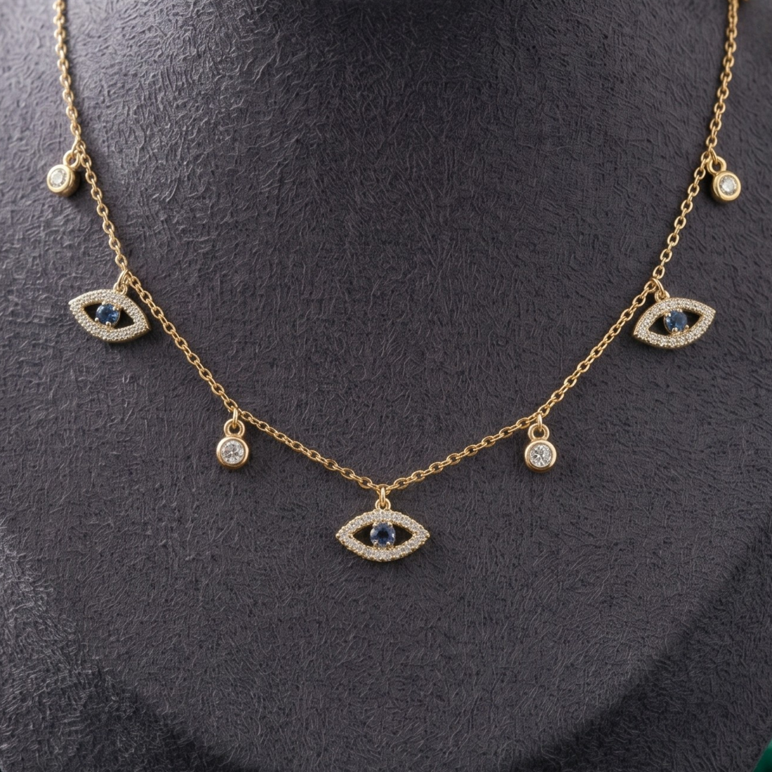 A gold chain necklace displayed on a dark gray mannequin bust, featuring three evil eye charms with dark blue centers and small clear stone accents.