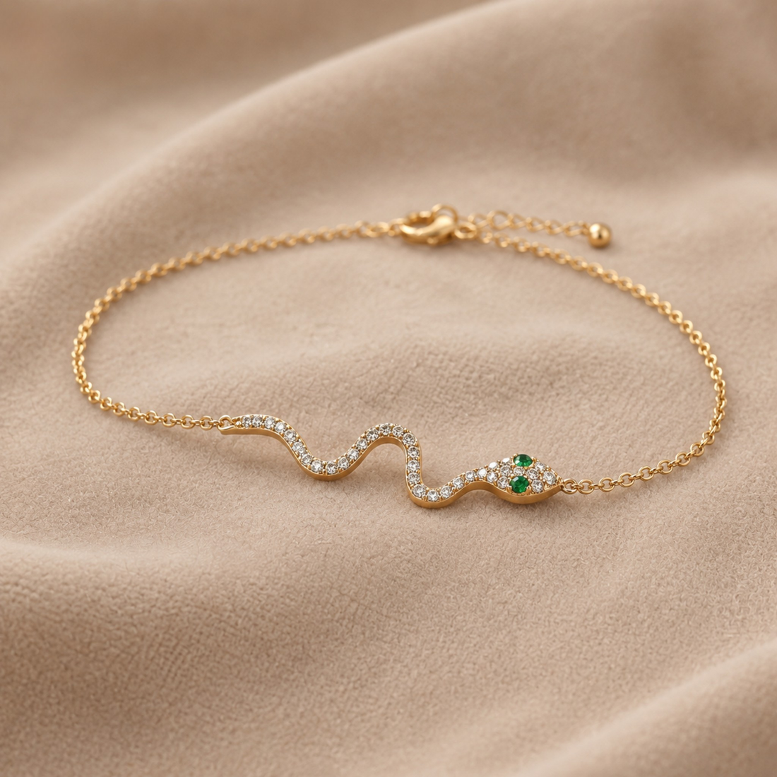 A delicate gold chain bracelet featuring a wavy snake charm paved with small clear stones and accented with a green emerald at its head, displayed on a soft beige fabric background.