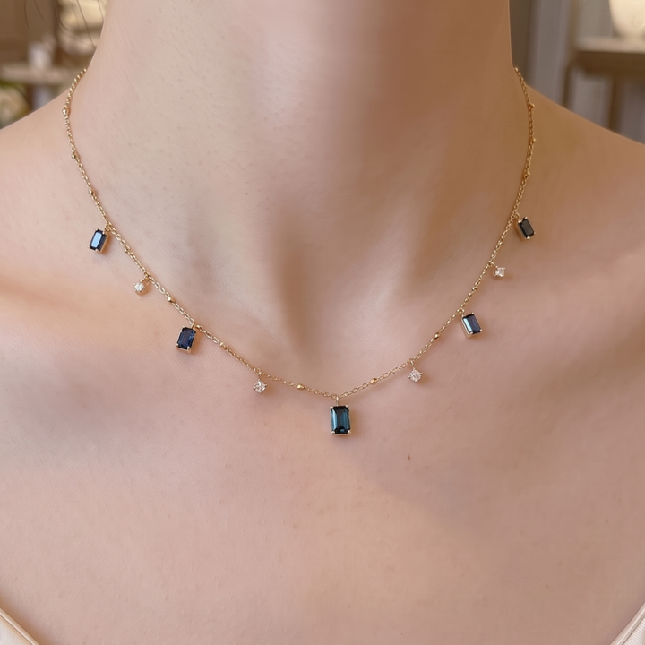 A delicate gold chain necklace with small dark blue rectangular stones and tiny clear accent stones, shown worn on a model's neck.