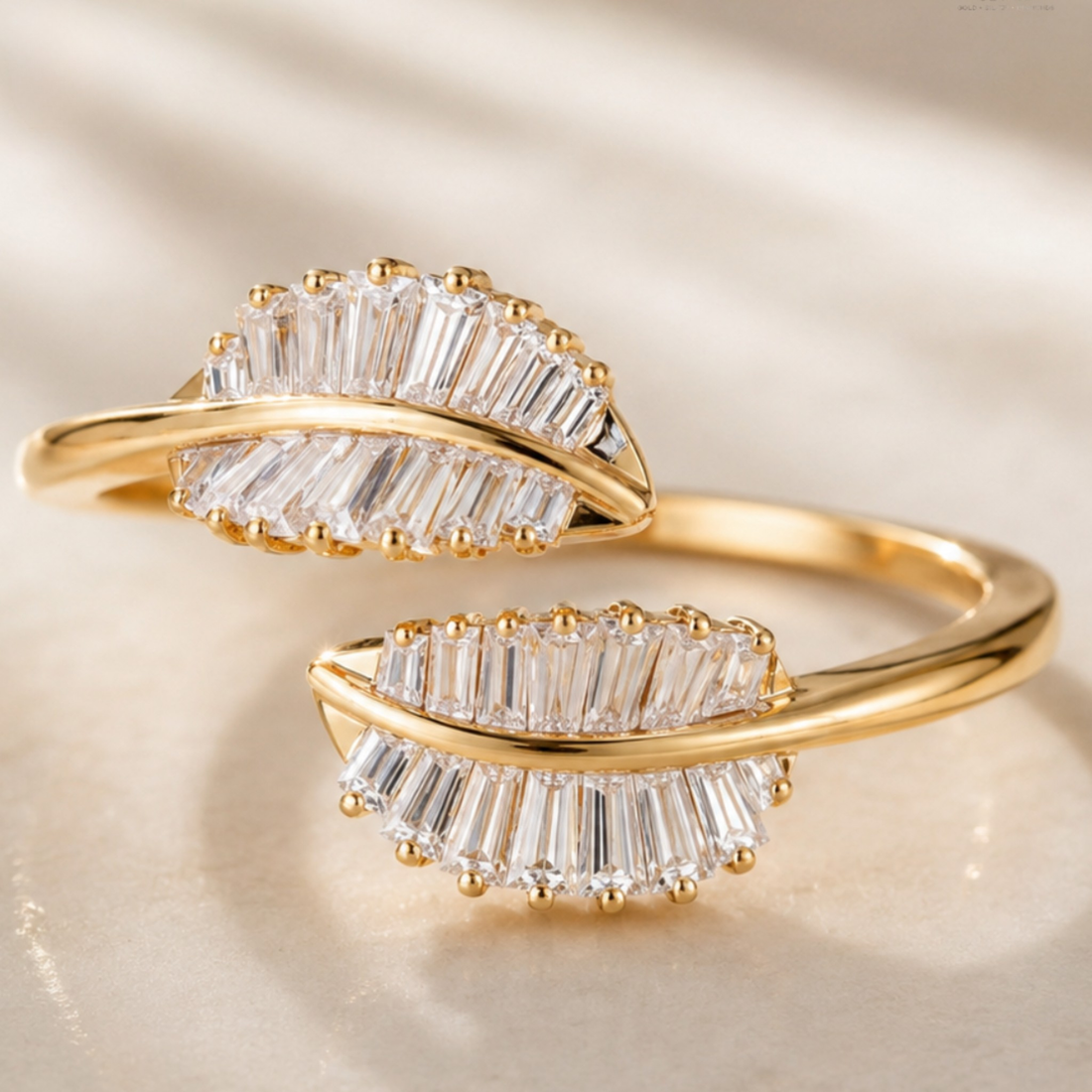 A gold ring designed in the shape of two overlapping leaves, each adorned with clear baguette-cut gemstones and small gold bead accents, set against a soft beige background.
