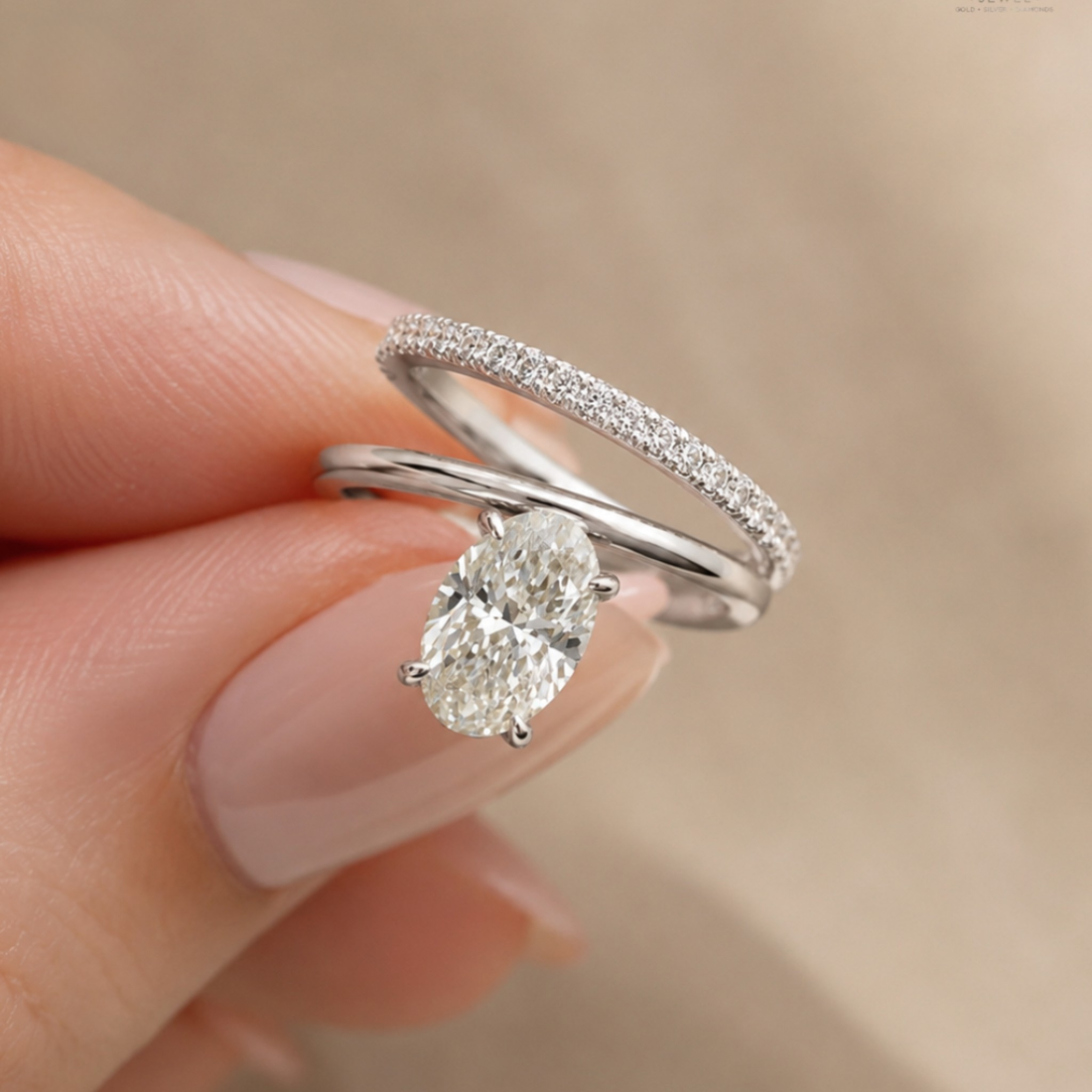 A close-up of a hand wearing a silver engagement ring with a large oval-cut center stone and a matching pavé band ring behind it, set against a soft beige background.