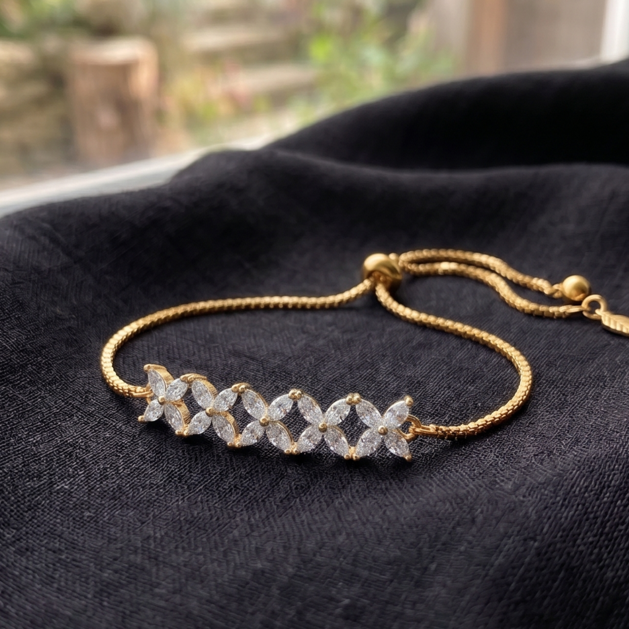 A gold-plated bracelet with a row of marquise-cut clear stones set in a gold chain, resting on a textured black fabric surface.