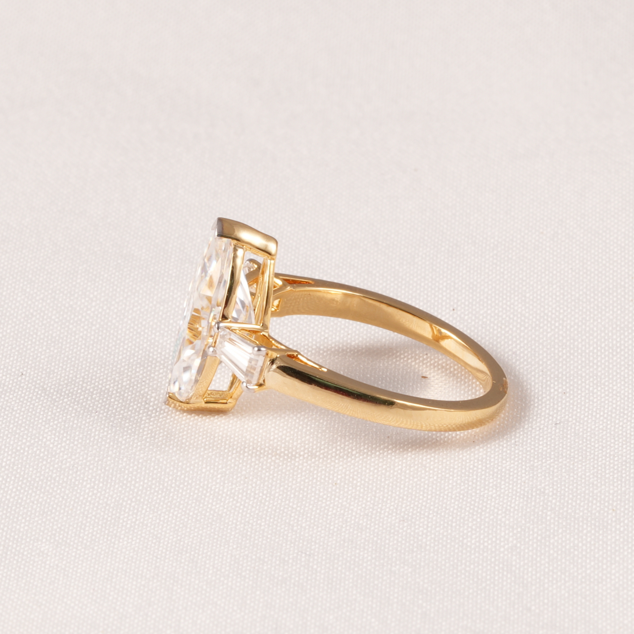 A gold ring with a large rectangular clear gemstone in a prong setting, photographed on a light beige textured fabric background.