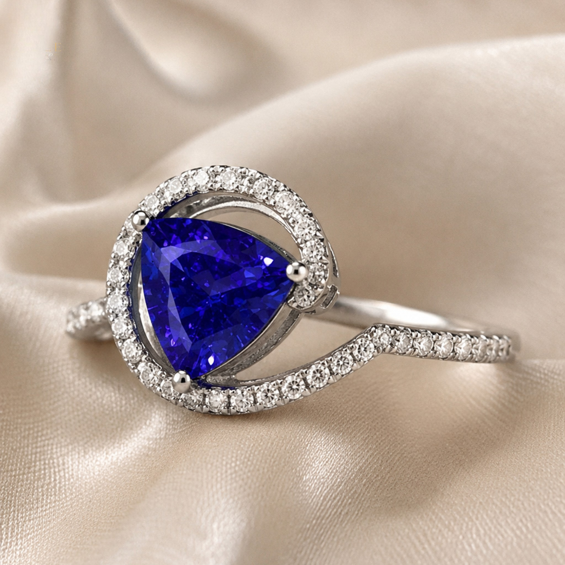 A silver ring featuring a large pear-cut blue sapphire center stone surrounded by a halo of small clear stones, with a curved pavé-set band, displayed on a beige satin fabric background.