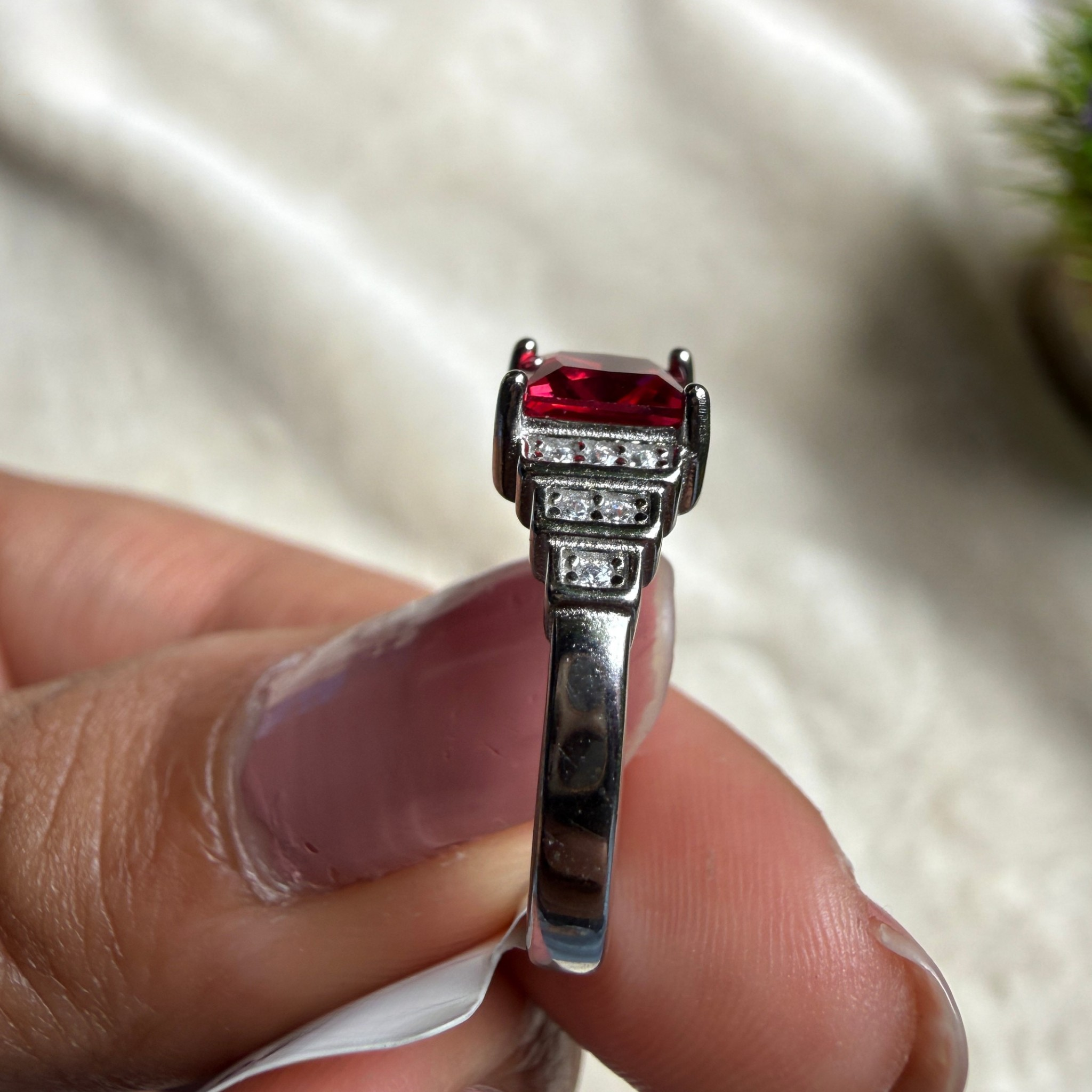 A close-up of a hand holding a silver ring with a rectangular red gemstone and small clear accent stones on a soft beige fabric background.