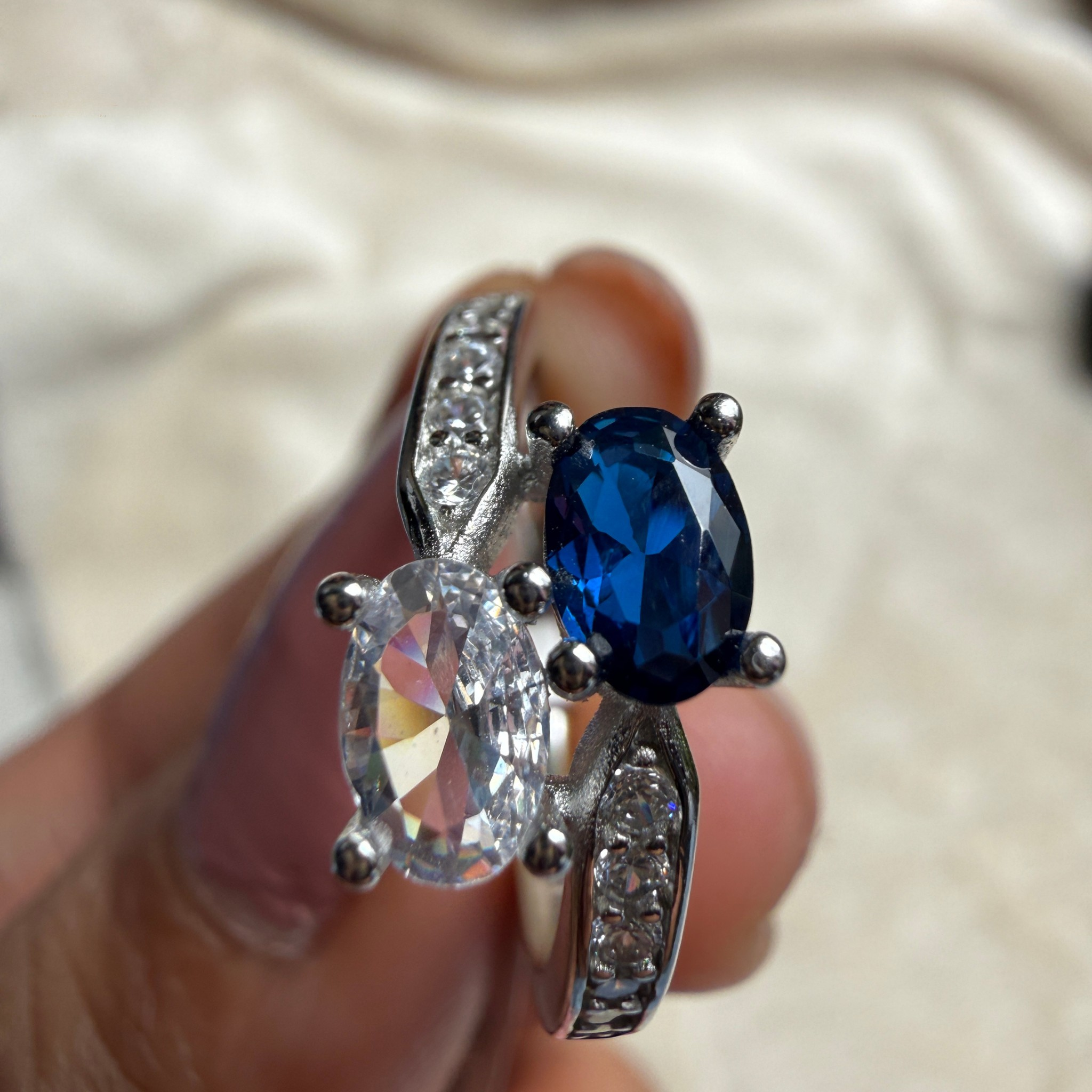 A close-up of a silver ring being held on a finger, featuring a large oval-cut blue gemstone and an oval clear gemstone, with small clear accent stones along the band.