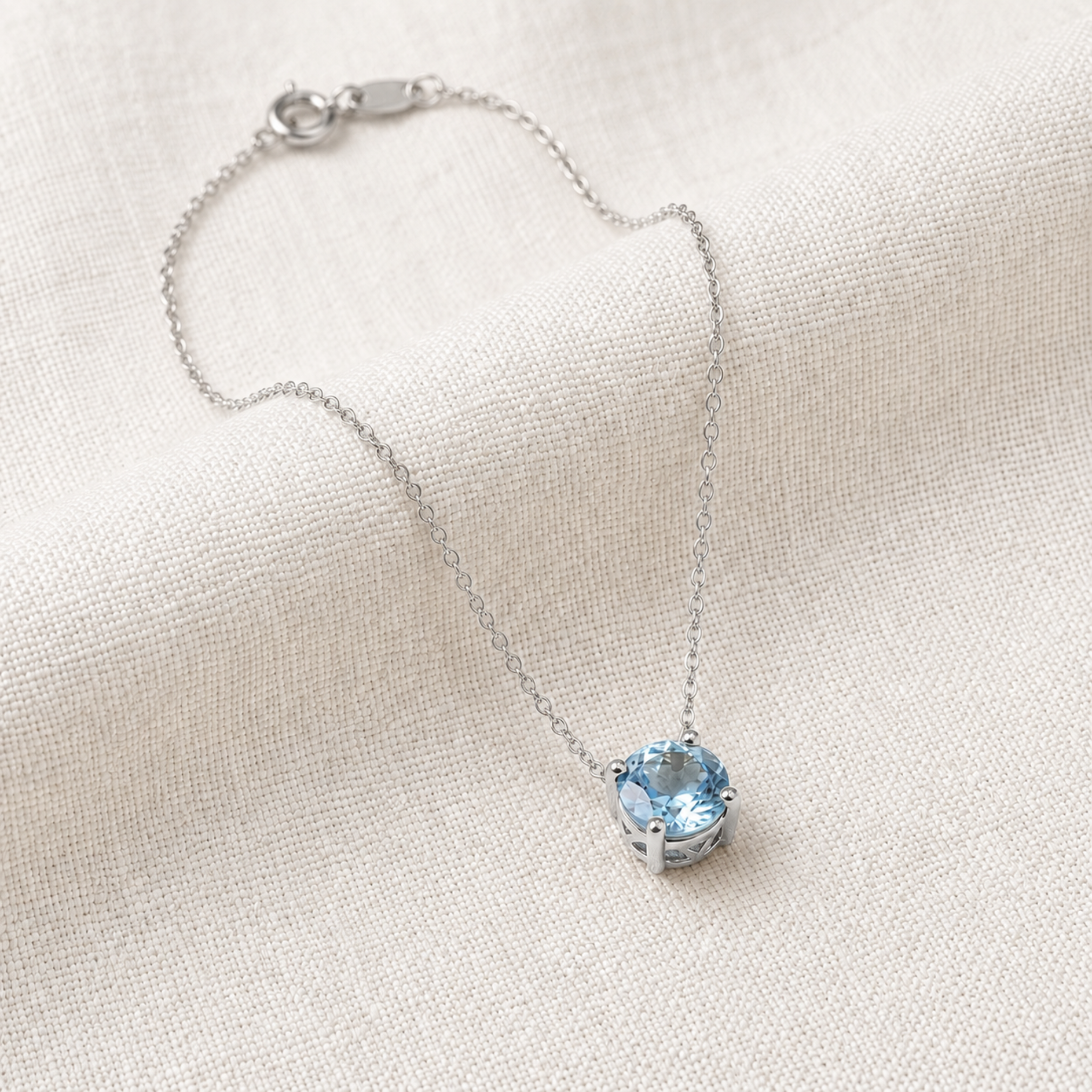 A delicate silver chain necklace with a small bezel-set light blue gemstone pendant, displayed on a textured off-white linen fabric background.