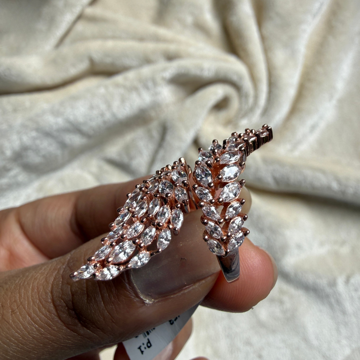 Rose gold wing-shaped ring with marquise-cut clear crystals, held between fingers against a draped cream fabric background.
