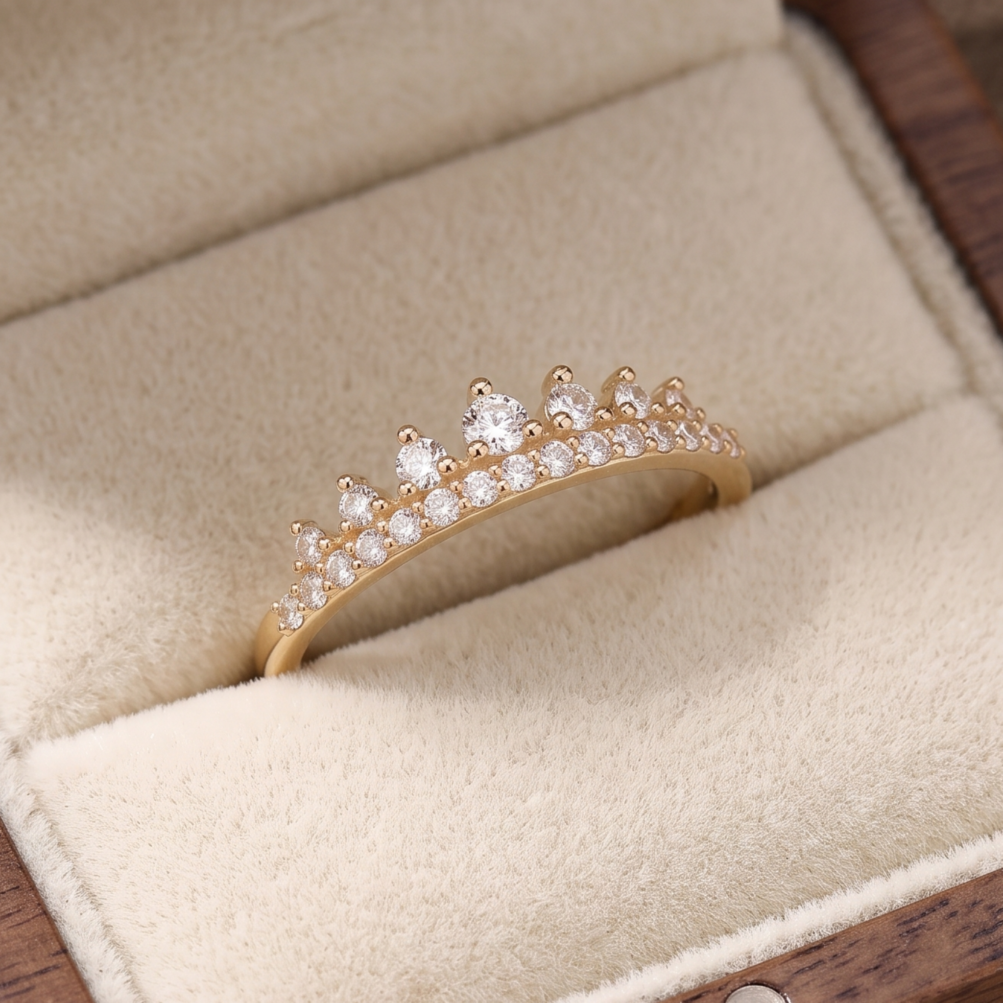 A gold crown-shaped ring set with clear gemstones, displayed inside a beige velvet jewelry box.