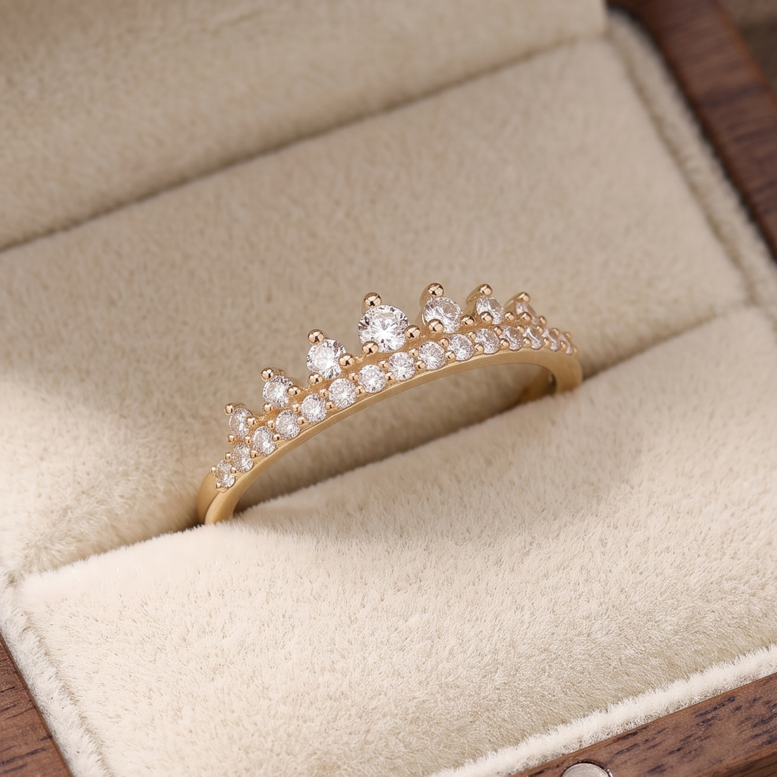 A gold crown-shaped ring set with clear gemstones, displayed inside a beige velvet jewelry box.