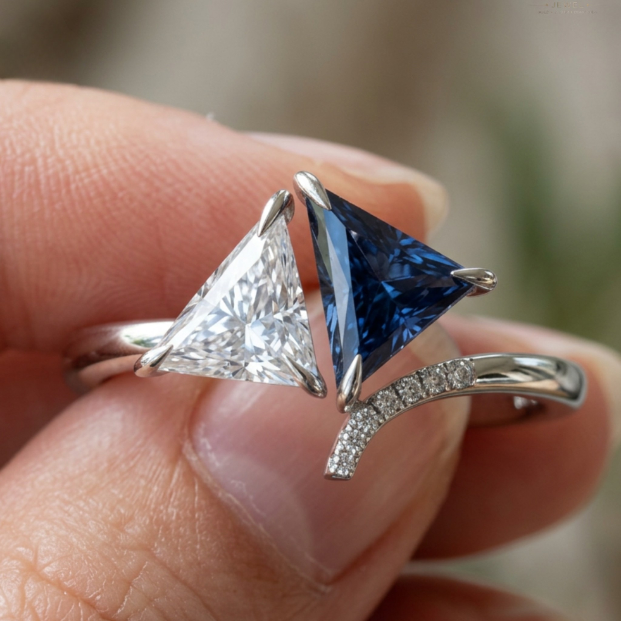 A close-up of a silver ring held between fingers, featuring two triangular gemstones: one clear and one deep blue, with a curved silver band accented by small pavé stones.