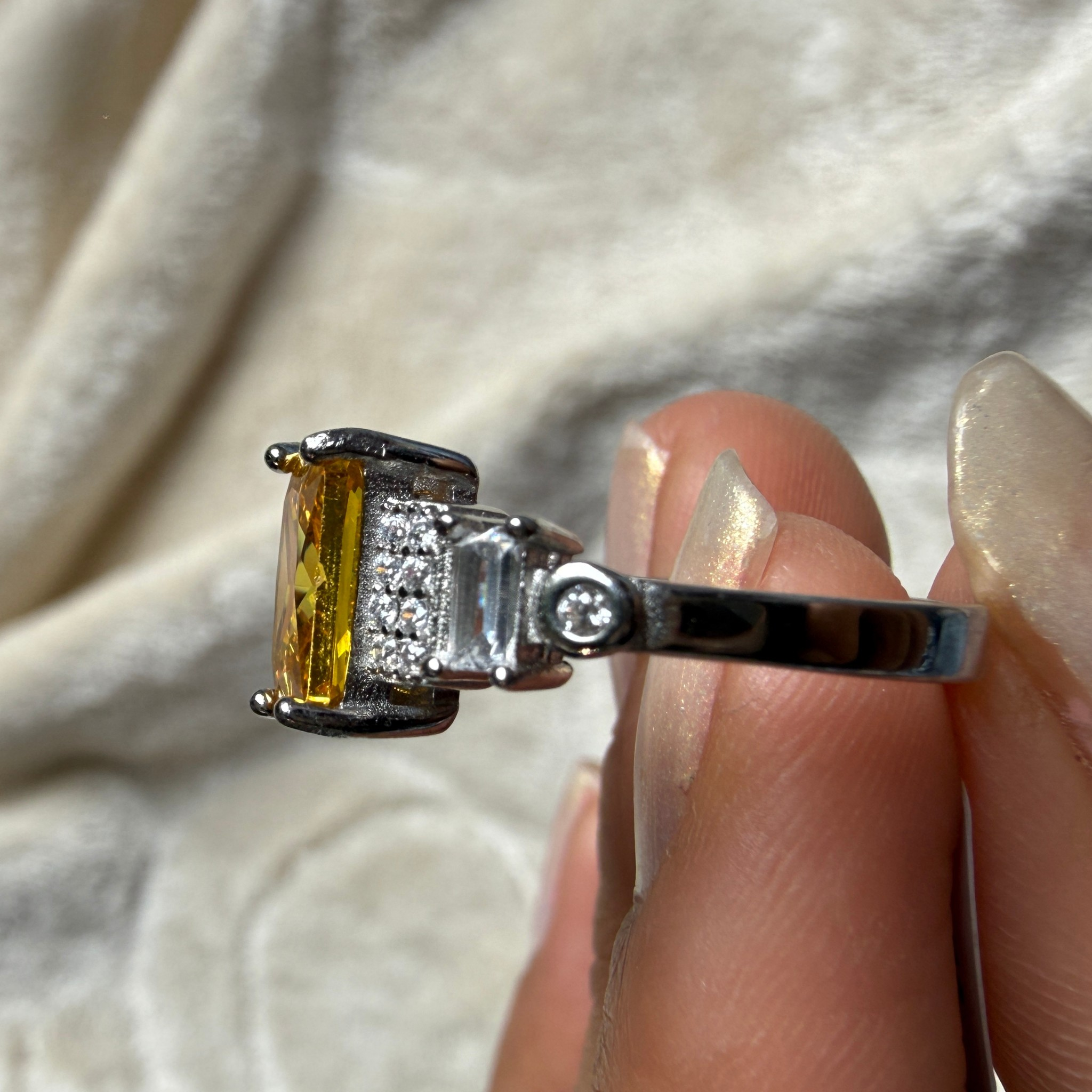 A silver ring with a rectangular yellow gemstone and small clear accent stones, held between fingers against a soft beige fabric background.