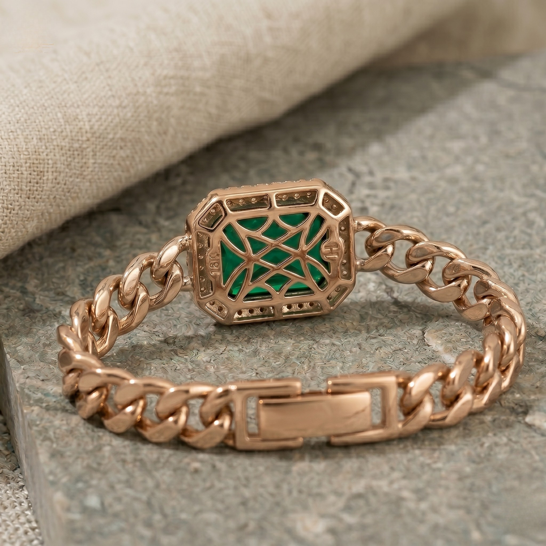 A rose gold chain bracelet with a central octagonal green and gold geometric inlay, displayed on a textured gray stone surface with a beige linen cloth in the background.