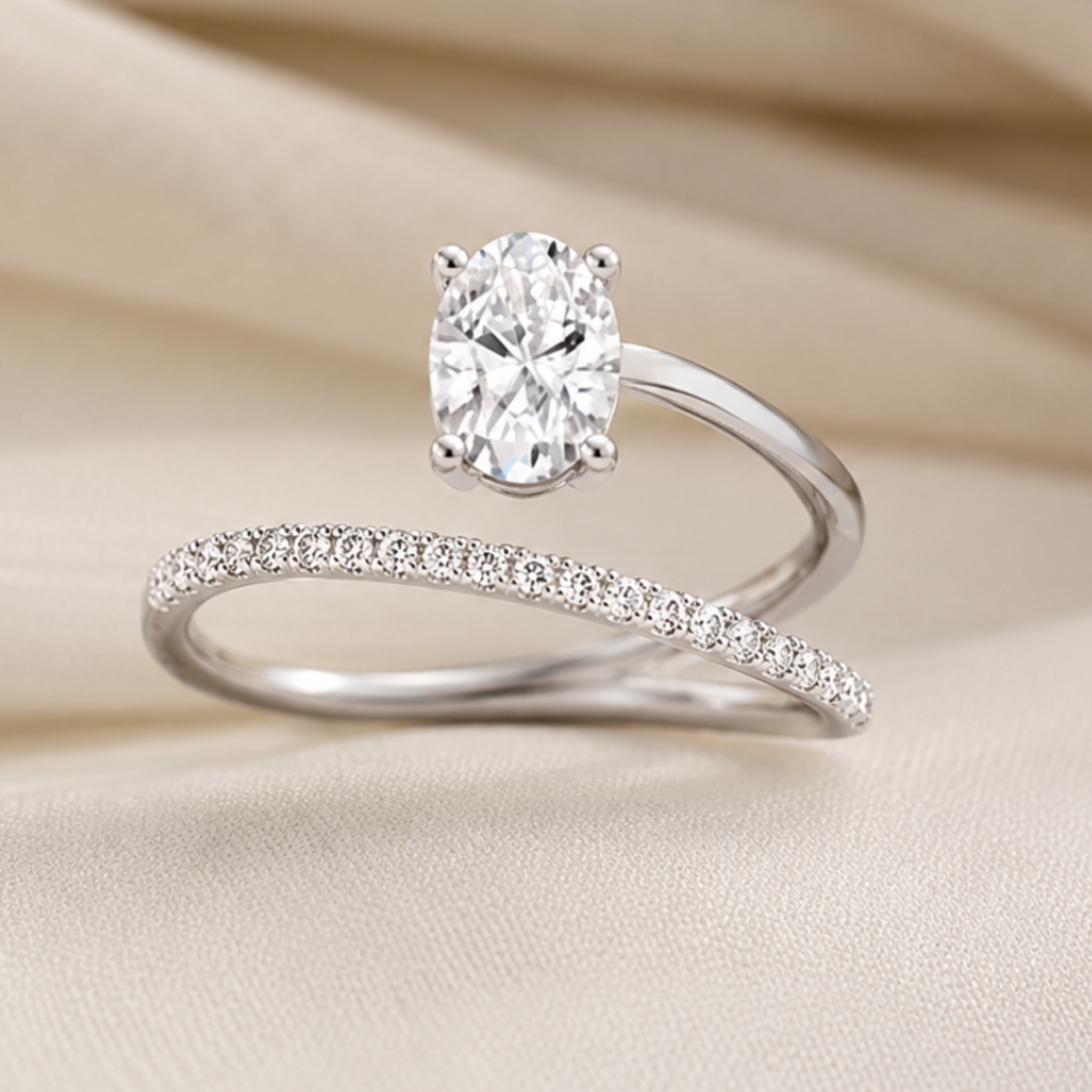 A silver engagement ring with a round brilliant-cut diamond is displayed on a draped beige fabric background, with a matching silver band featuring a row of small pavé diamonds positioned underneath.