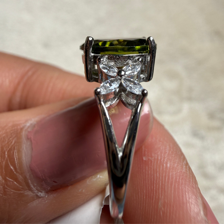 A close-up of a silver ring held between fingers, featuring a rectangular green gemstone at the top and a flower-shaped cluster of clear stones below.