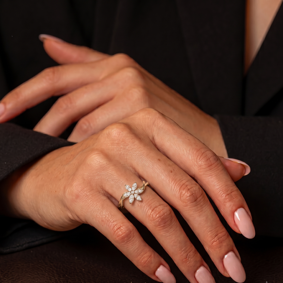 A close-up of a person's hands wearing a delicate gold ring with a small flower-shaped cluster of clear stones, styled with a black blazer and light pink manicured nails.