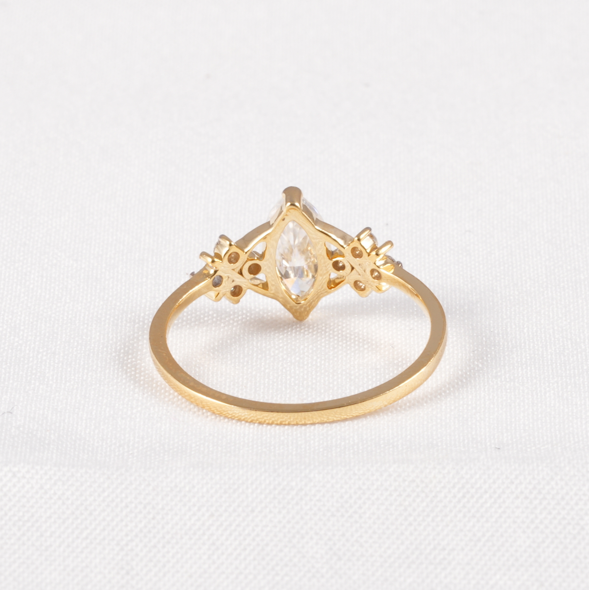 A gold ring featuring a marquise-cut clear gemstone in a decorative diamond-shaped setting with small side accents, shown on a textured white background.