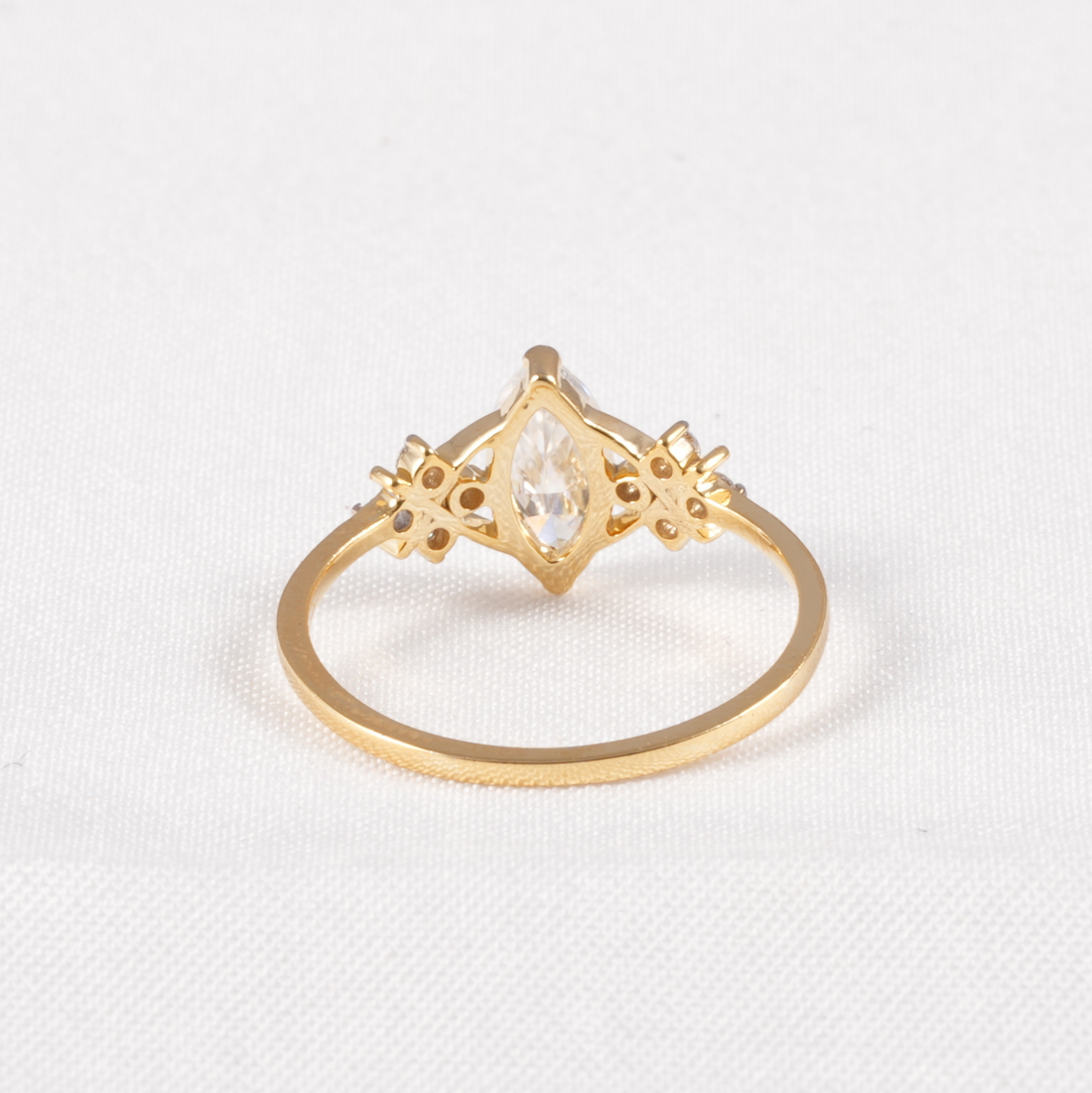 A gold ring featuring a marquise-cut clear gemstone in a decorative diamond-shaped setting with small side accents, shown on a textured white background.