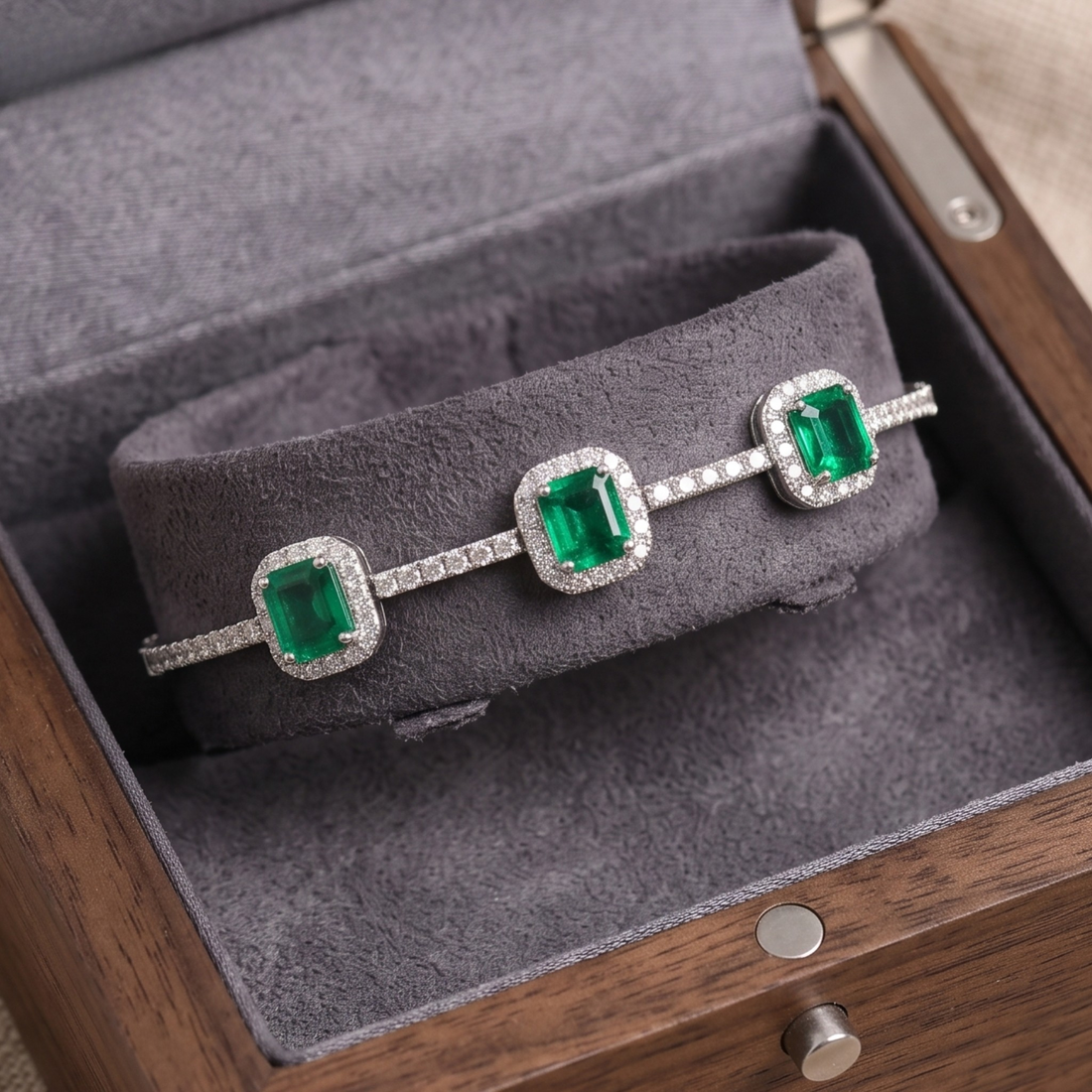 A silver-toned bracelet with three emerald-cut green gemstones and pavé-set clear stones, displayed on a gray velvet cushion inside a wooden presentation box.