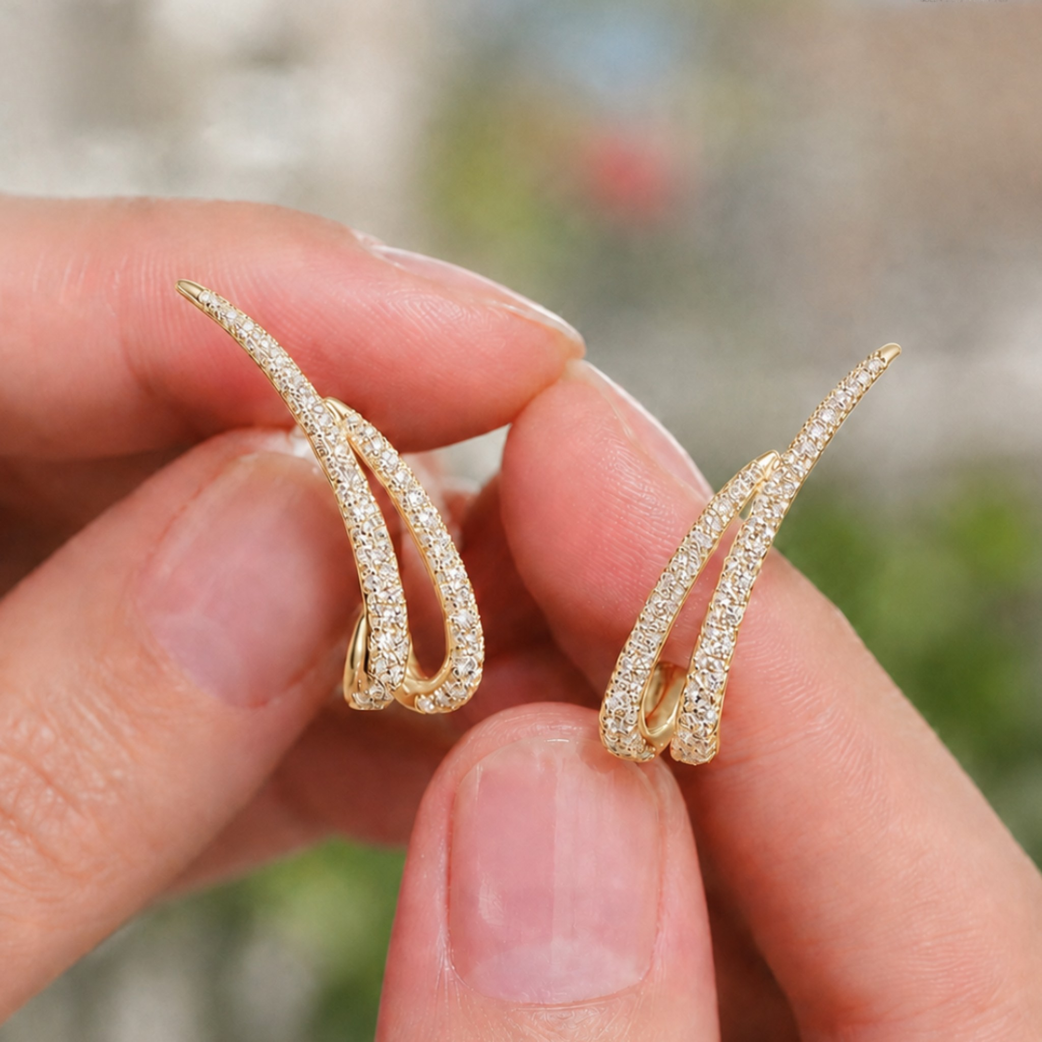 A pair of gold huggie hoop earrings paved with small clear stones, held between two fingers against a blurred outdoor background.