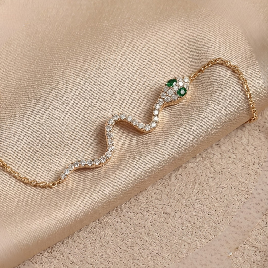 A gold snake necklace with clear pavé stones and green emerald eyes, displayed on a beige satin fabric background.