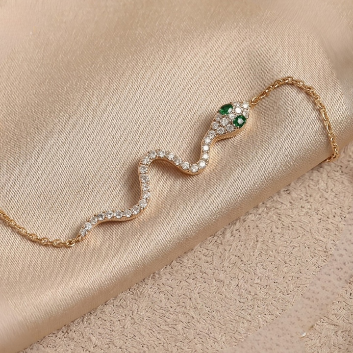 A gold snake necklace with clear pavé stones and green emerald eyes, displayed on a beige satin fabric background.