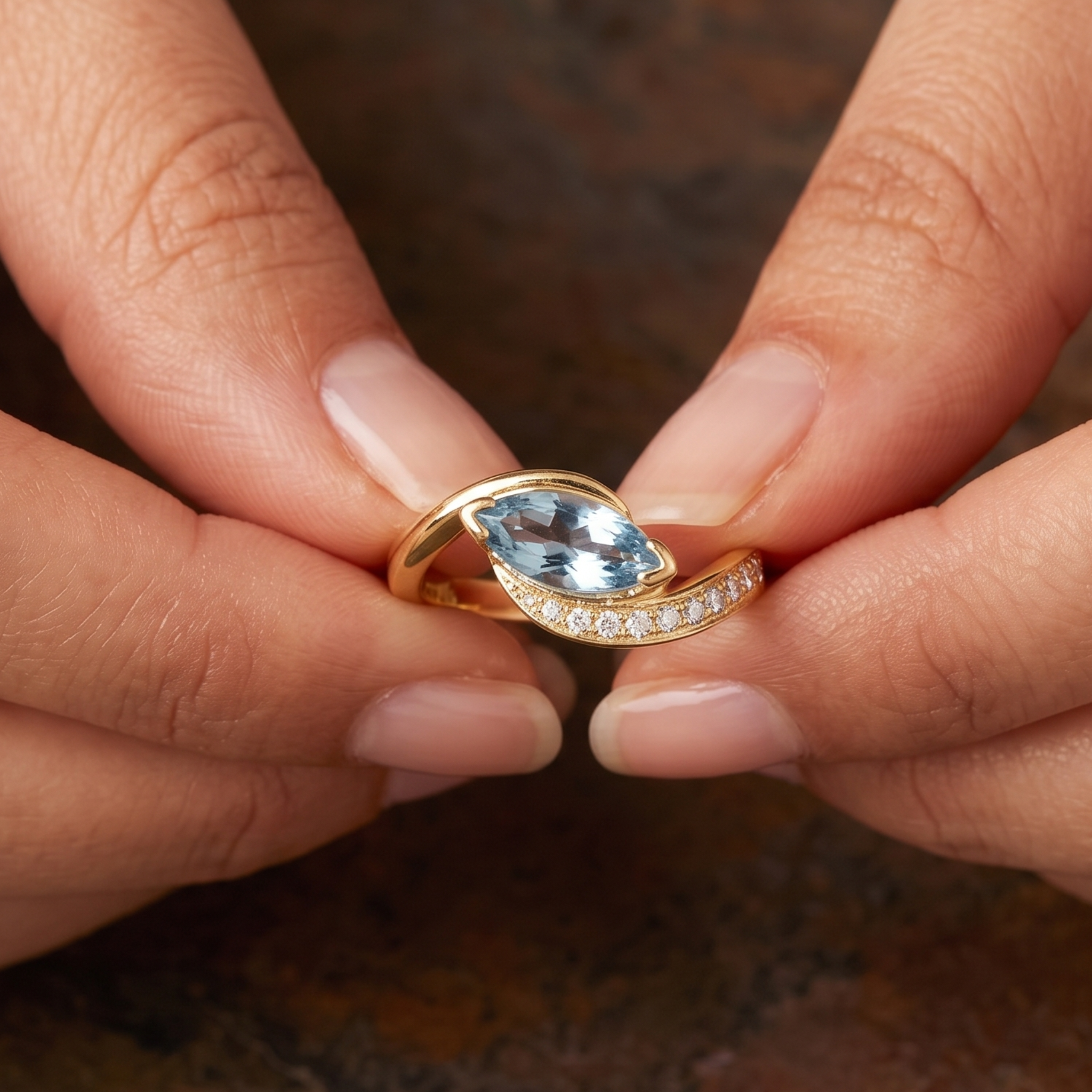 A close-up of a gold ring with a marquise-cut light blue gemstone and a curved row of small clear stones, held between two fingers against a dark brown textured background.