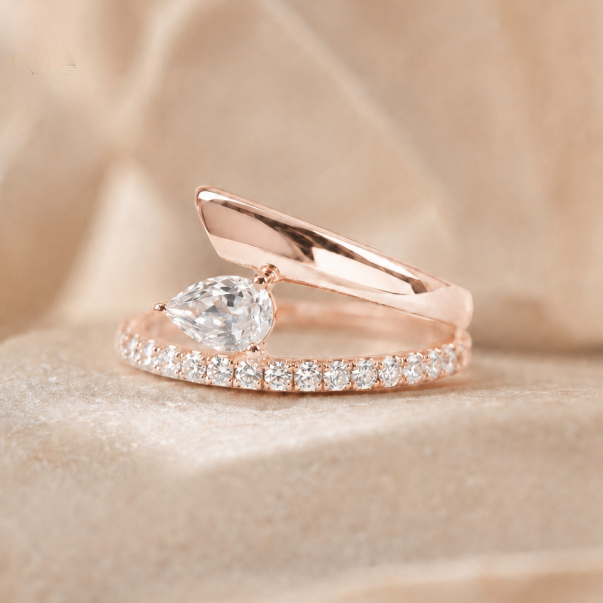 A rose gold ring featuring a pear-cut clear center stone and a pavé band, displayed on a soft beige fabric background.